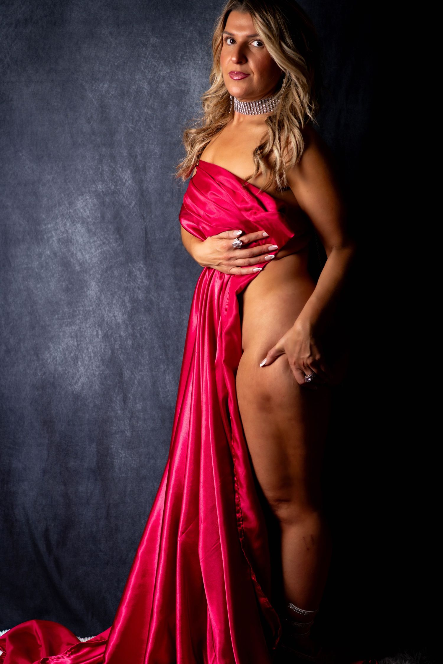Blonde woman posing in a flowing red satin gown against a dark studio backdrop.