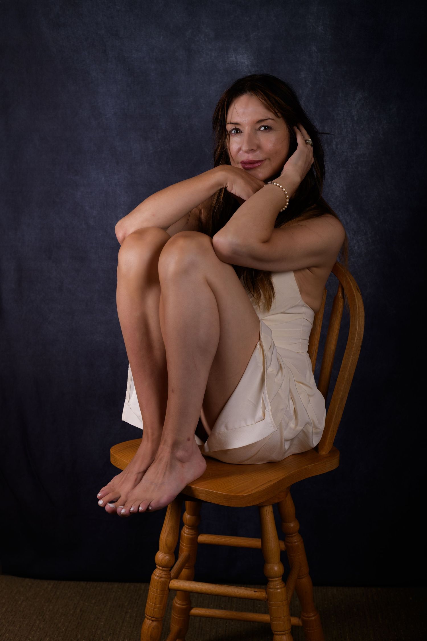 Woman in white dress sitting barefoot on wooden chair against dark blue background, posing gracefully.