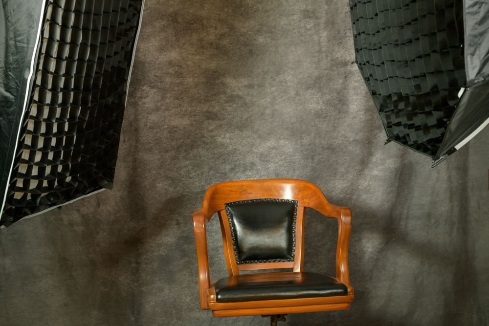 Vintage wooden chair with black leather seat cushion positioned against gray studio backdrop with lighting equipment.
