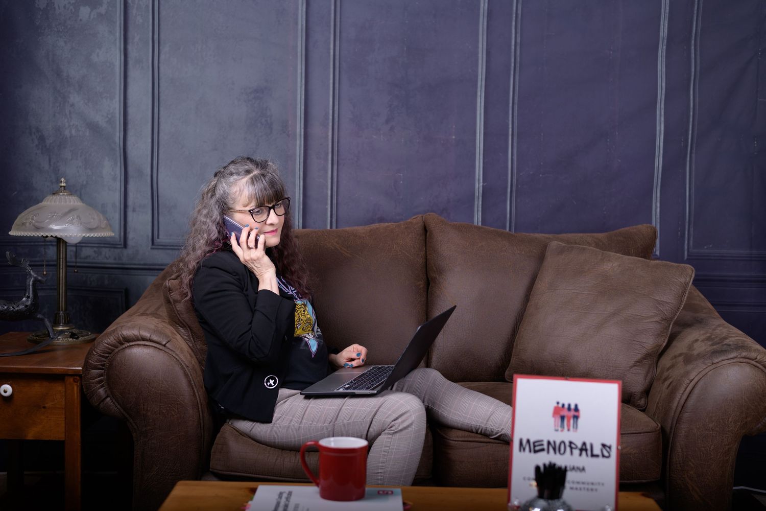 Woman sitting on brown leather sofa, talking on phone while using laptop, with red cup on coffee table nearby.