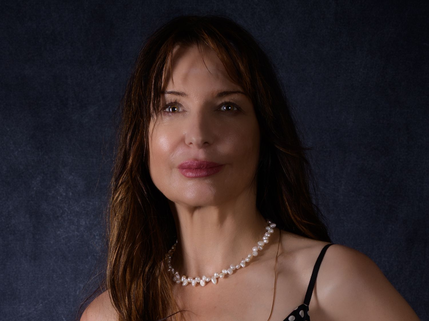 Elegant woman with long brown hair wearing a pearl necklace and black top poses for a portrait photo.