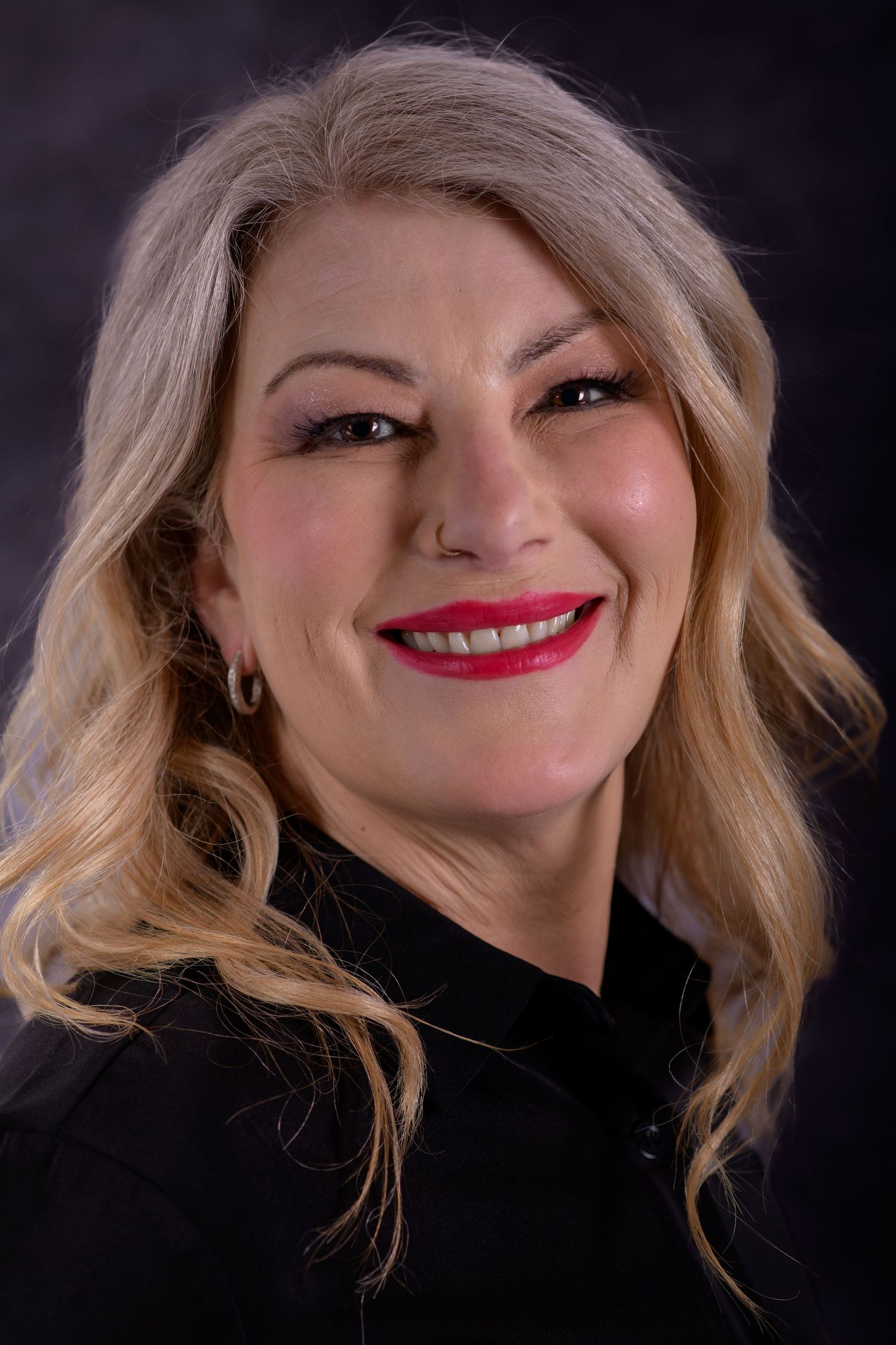 Smiling blonde woman with red lips wearing a black top against a dark studio background.