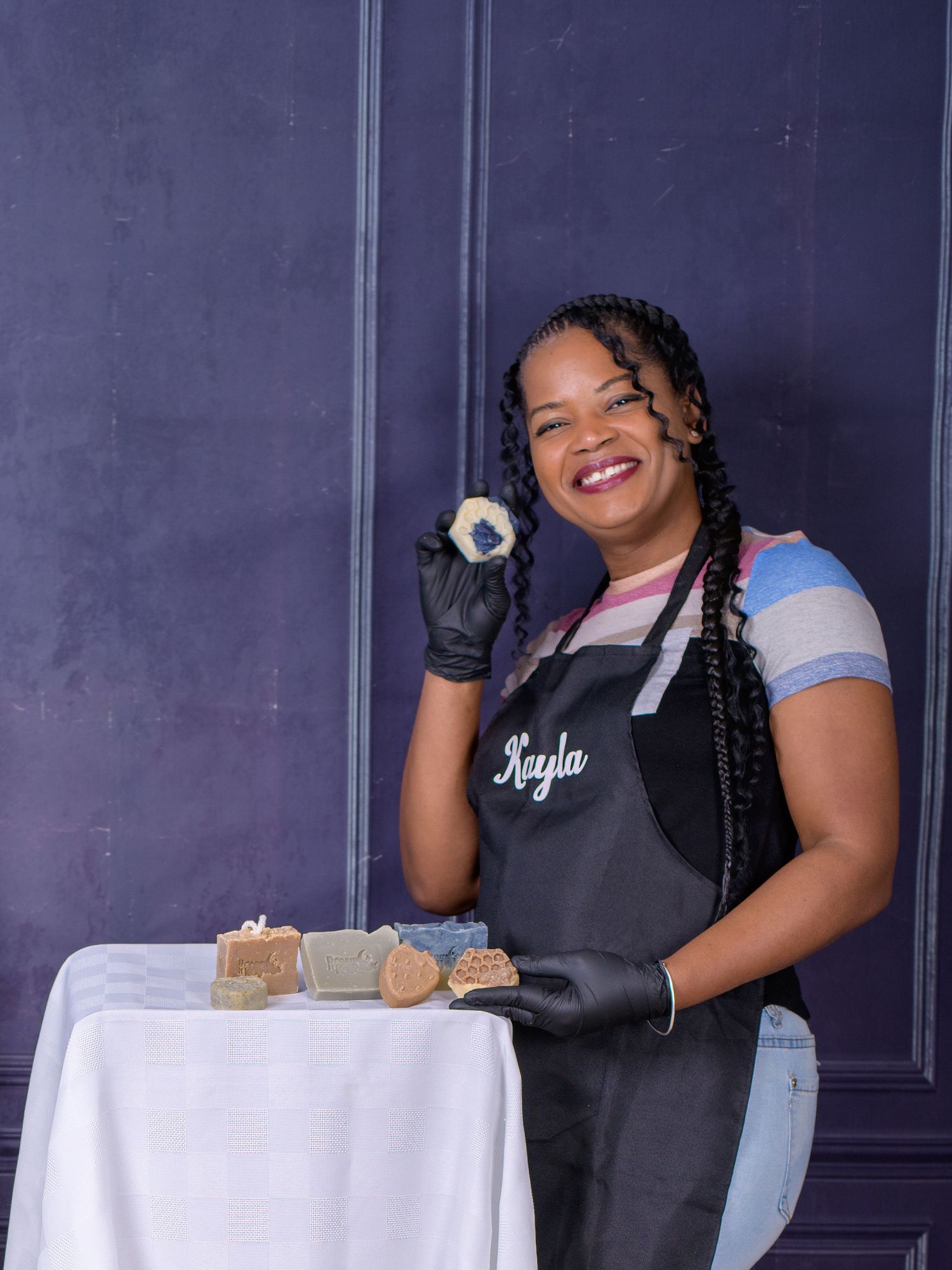 Woman named Kayla in black apron and gloves displays handmade soaps at a white table against a dark blue wall.