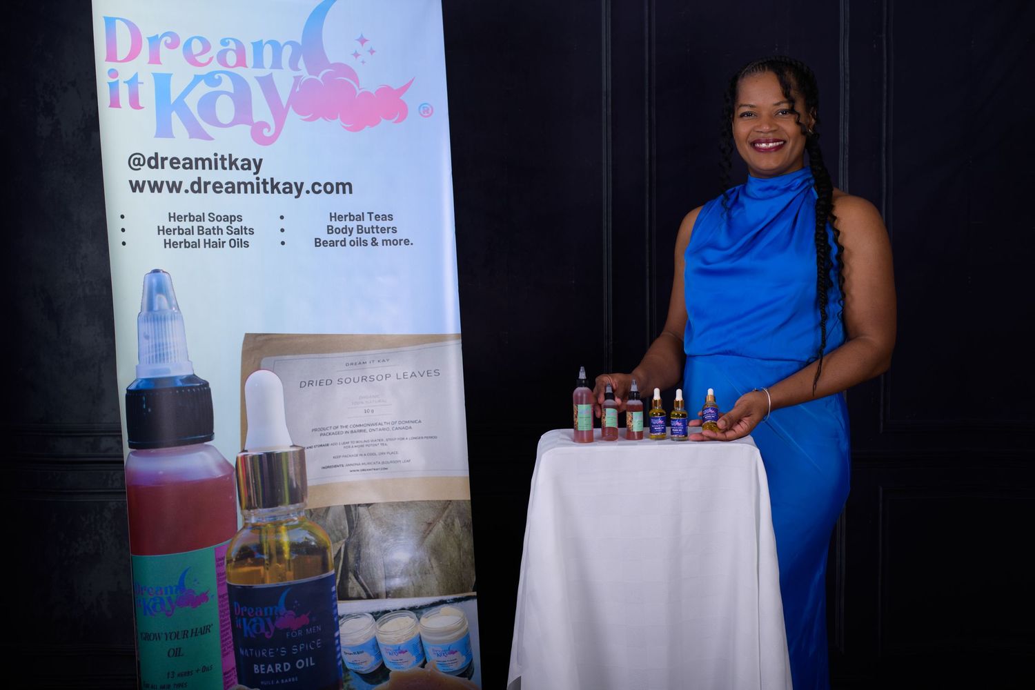 Woman in blue dress stands beside Dream It Kay herbal products display table at a vendor event.