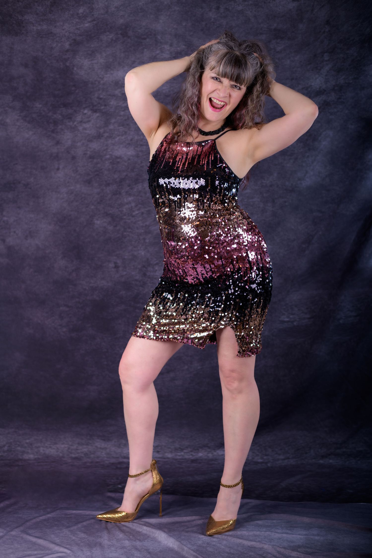 Woman in sequined multicolor mini dress and gold heels posing playfully with hands in hair on gray background.