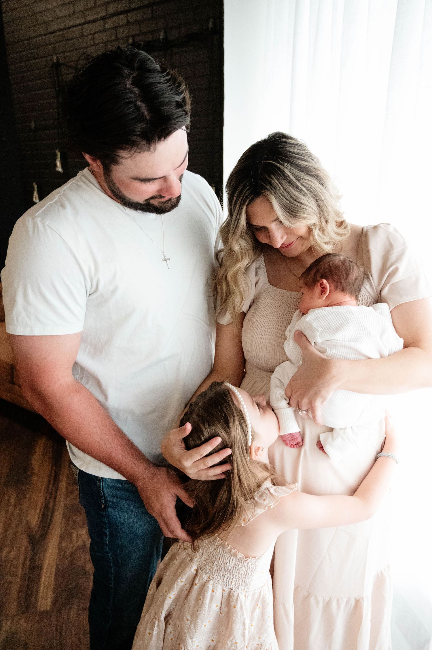 A family of four gazes lovingly at their newborn baby, with parents and young daughter gathered together near a bright window.