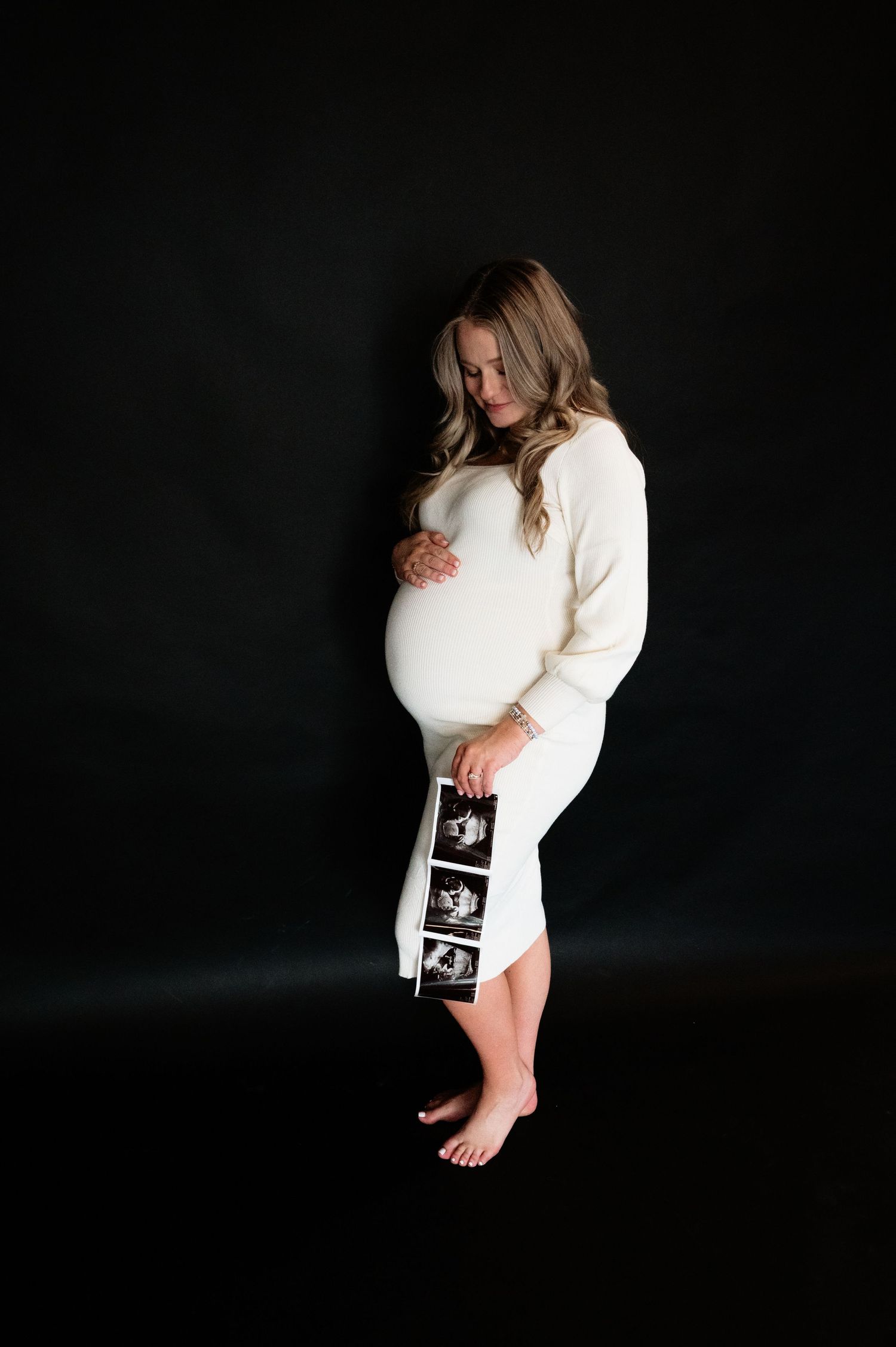 Expectant mother in white dress holds ultrasound images in dramatic black studio setting.