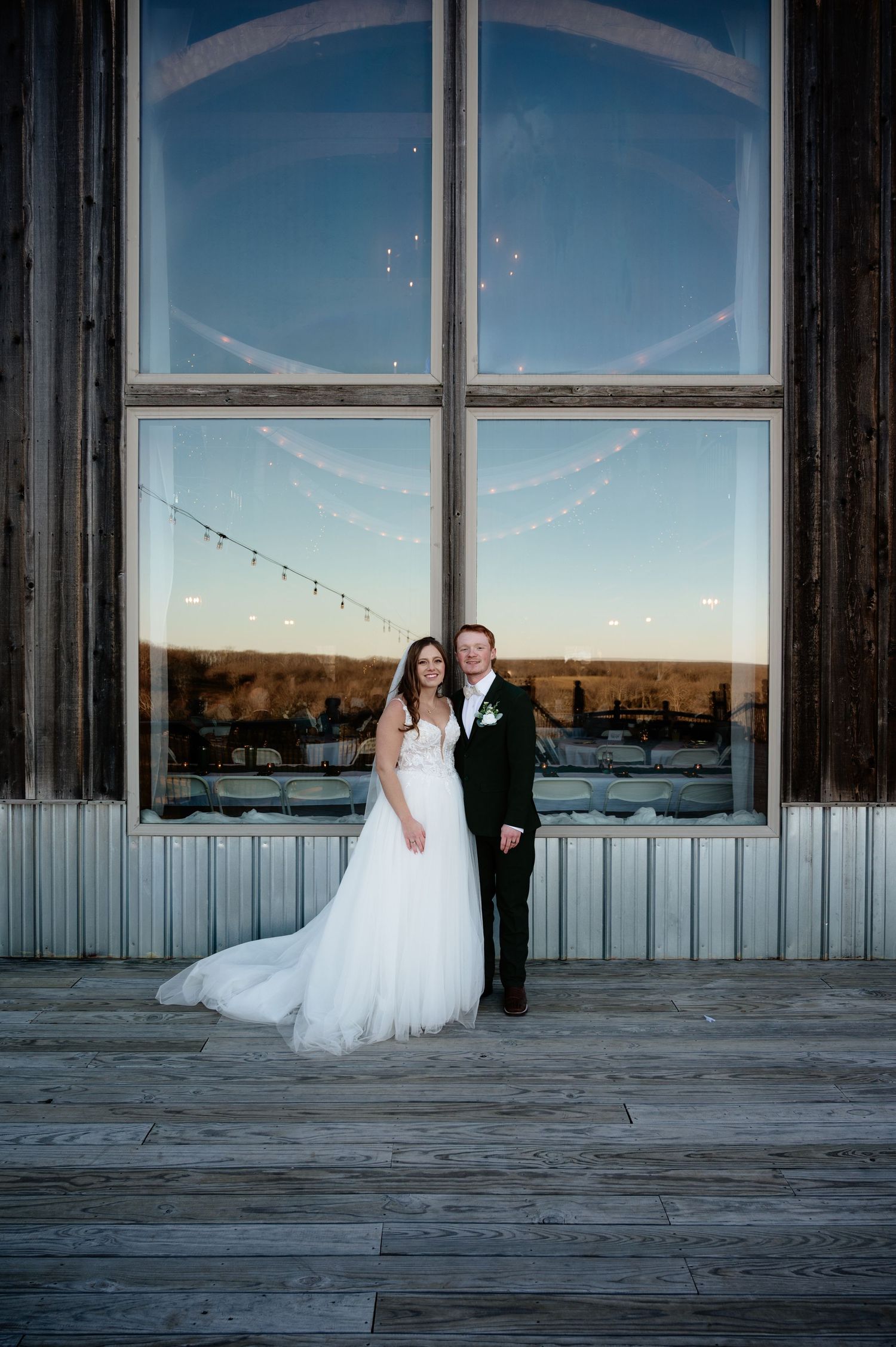 Bride in white gown and groom in dark suit pose before large window reflecting golden sunset at rustic wedding venue.