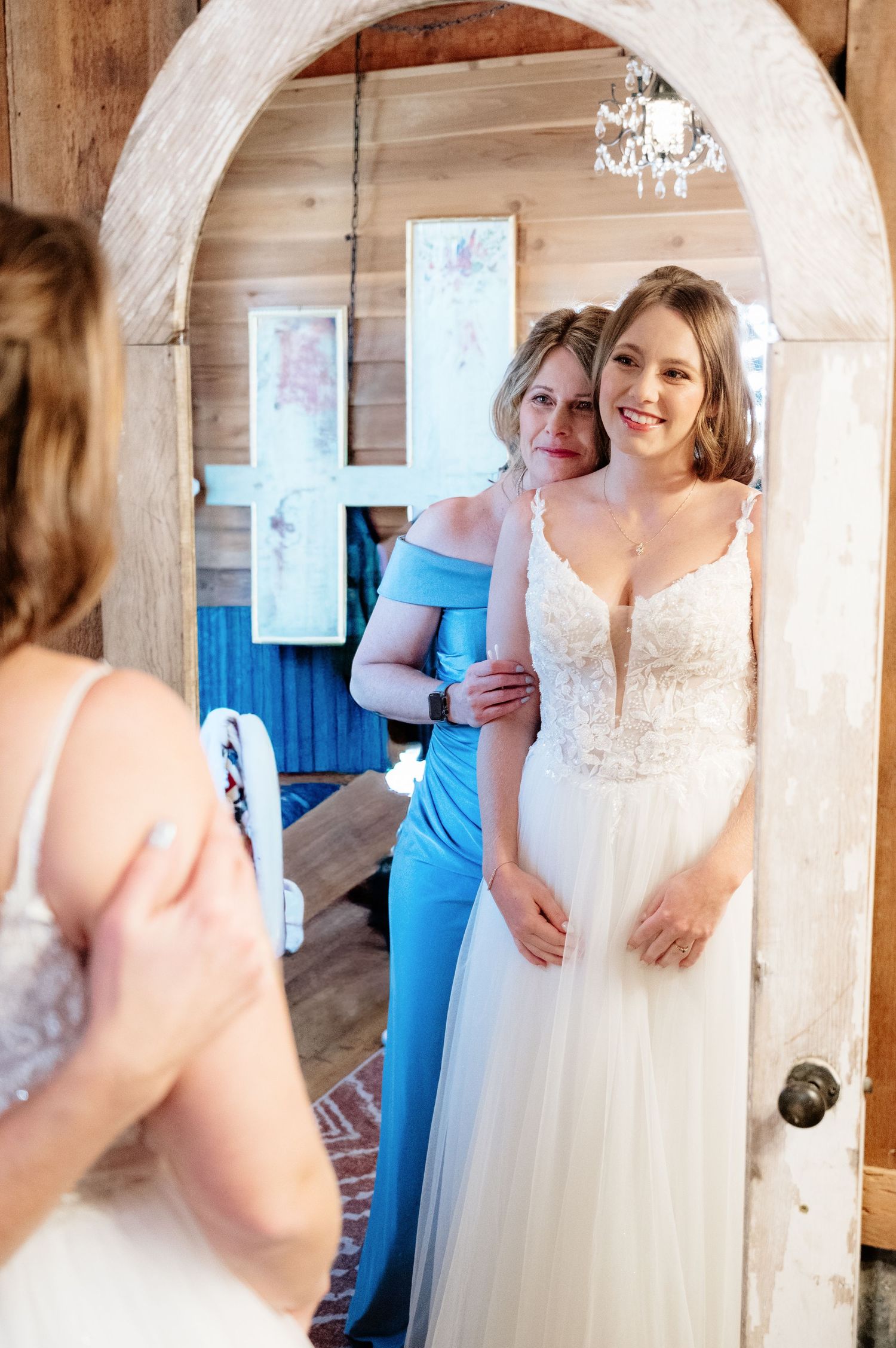 Bride in lace wedding gown smiles at mirror reflection while woman in blue dress helps her prepare for wedding day.