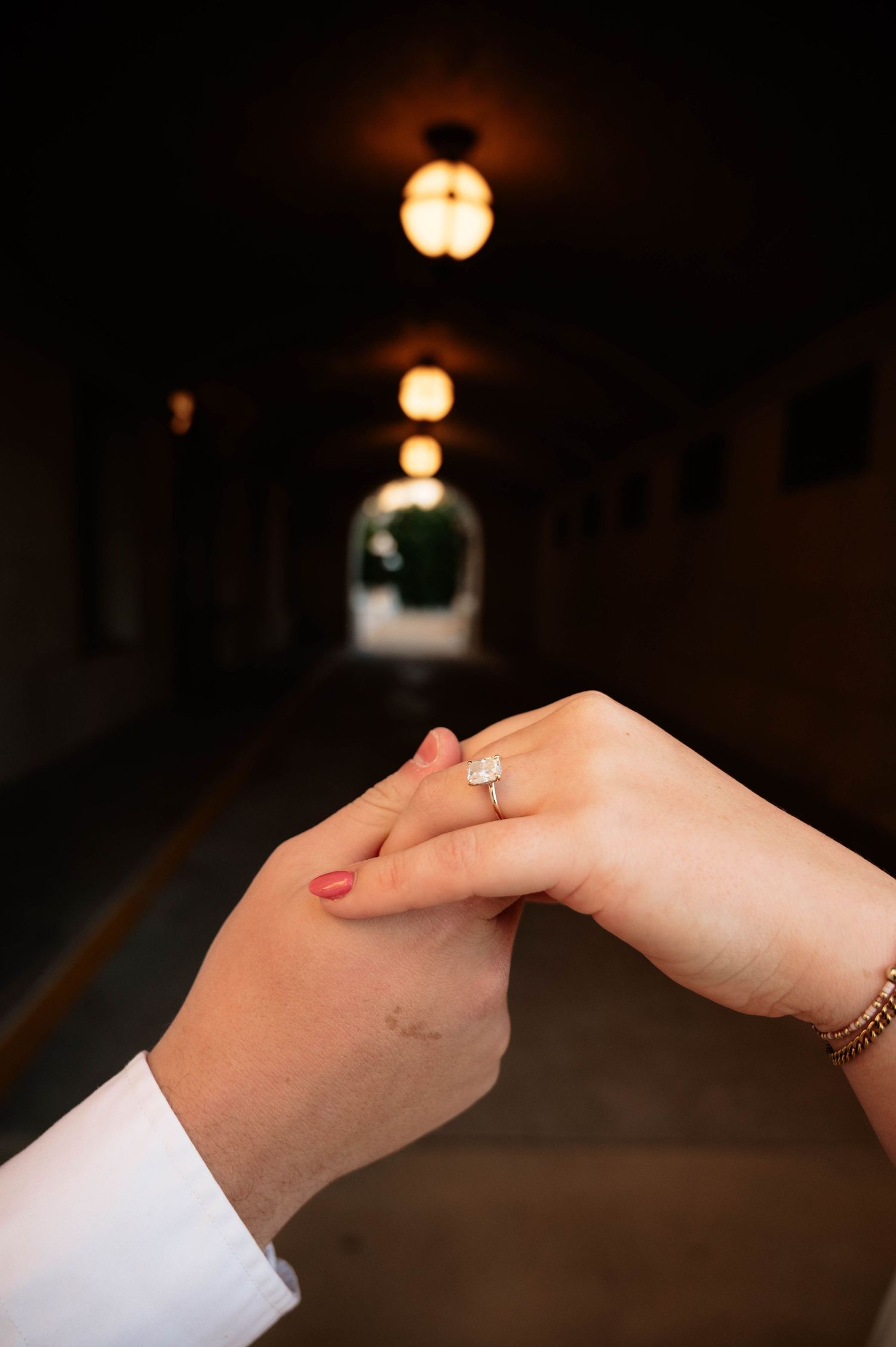 Two hands holding in a tunnel with warm lights, showcasing a sparkling diamond engagement ring on the woman's finger.