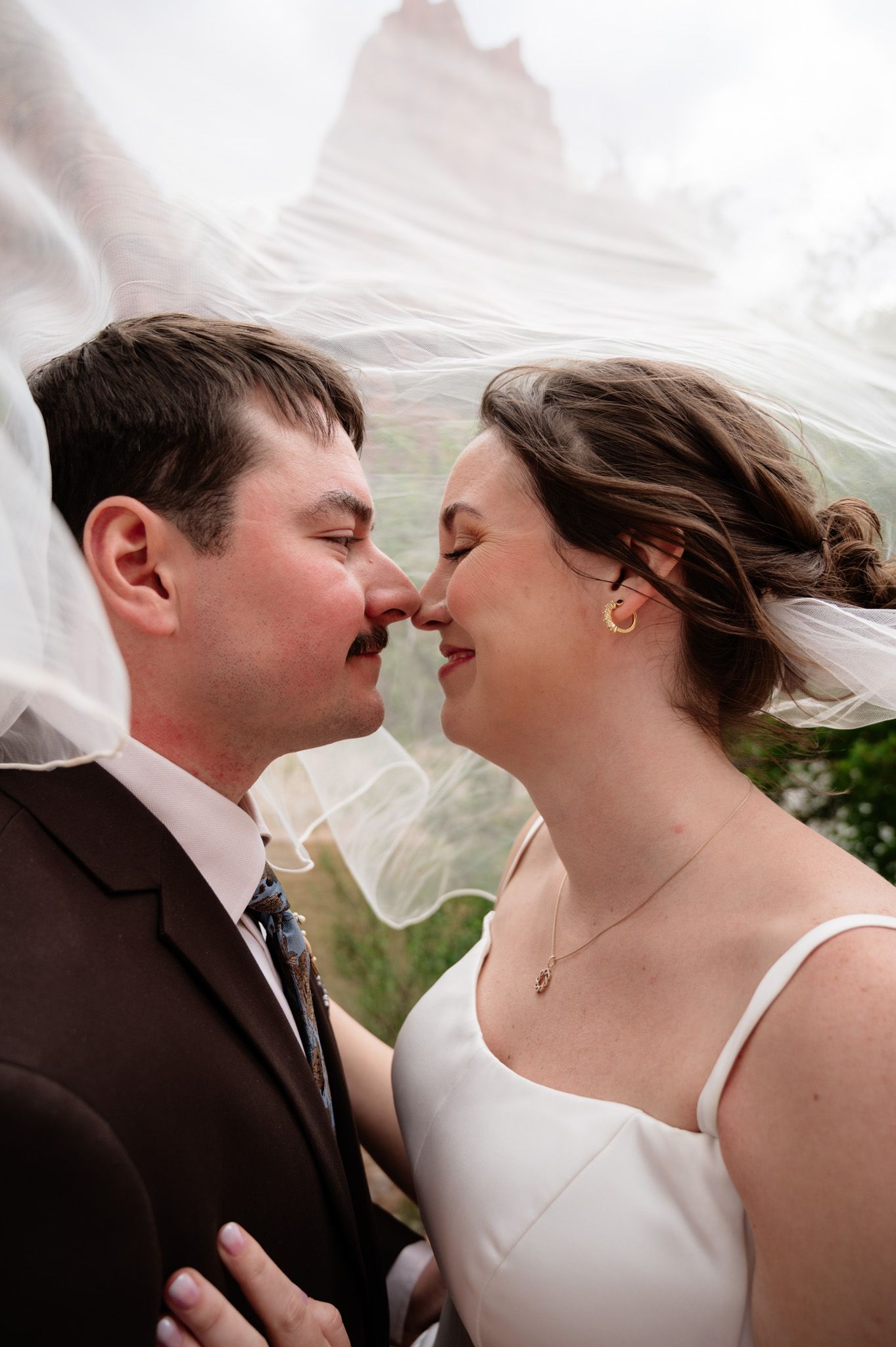 Bride and groom share an intimate nose-to-nose moment under a flowing veil with red rock formations in background.
