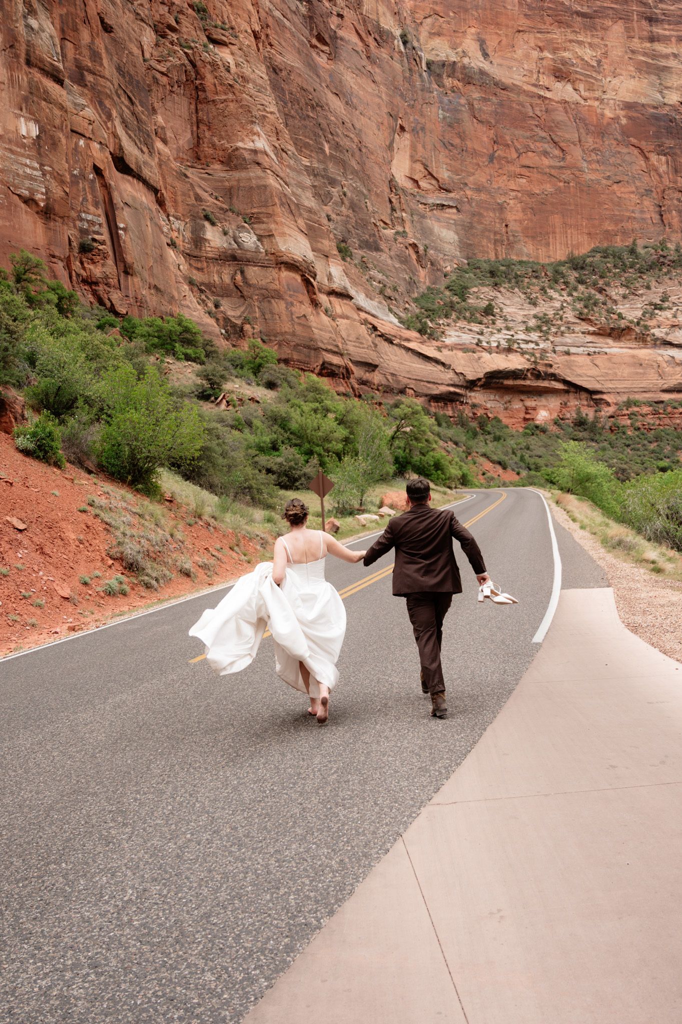 Bride and groom holding hands running down a scenic road in Zion National Park with red rock canyon walls.