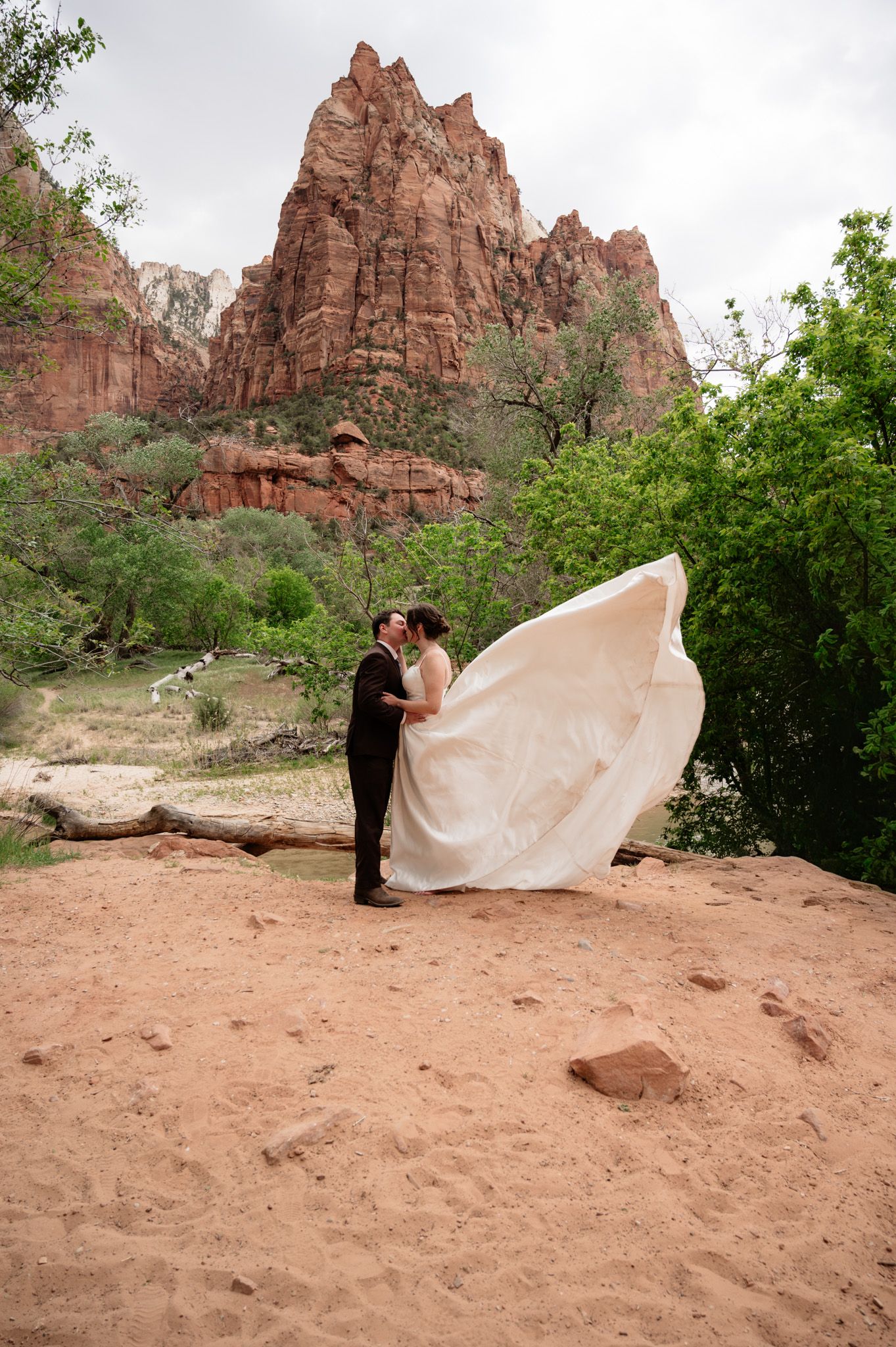 Couple kissing in wedding attire at Zion National Park, bride's dress flowing in wind with red rock formations behind.