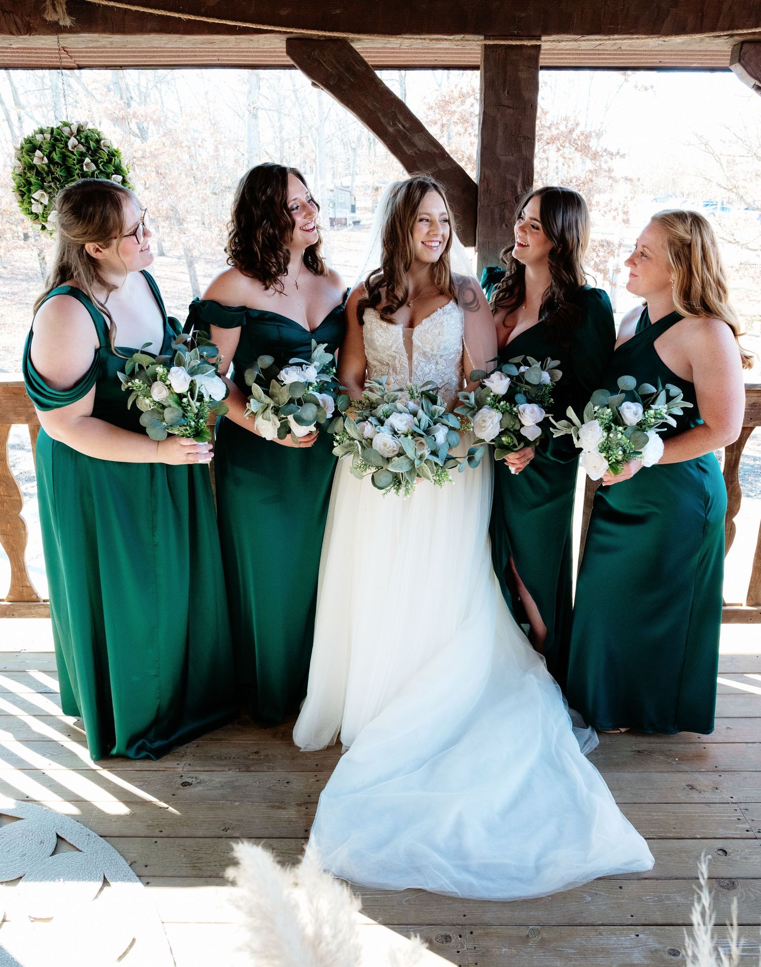 Wedding party poses in emerald green dresses and white gown under rustic covered area.
