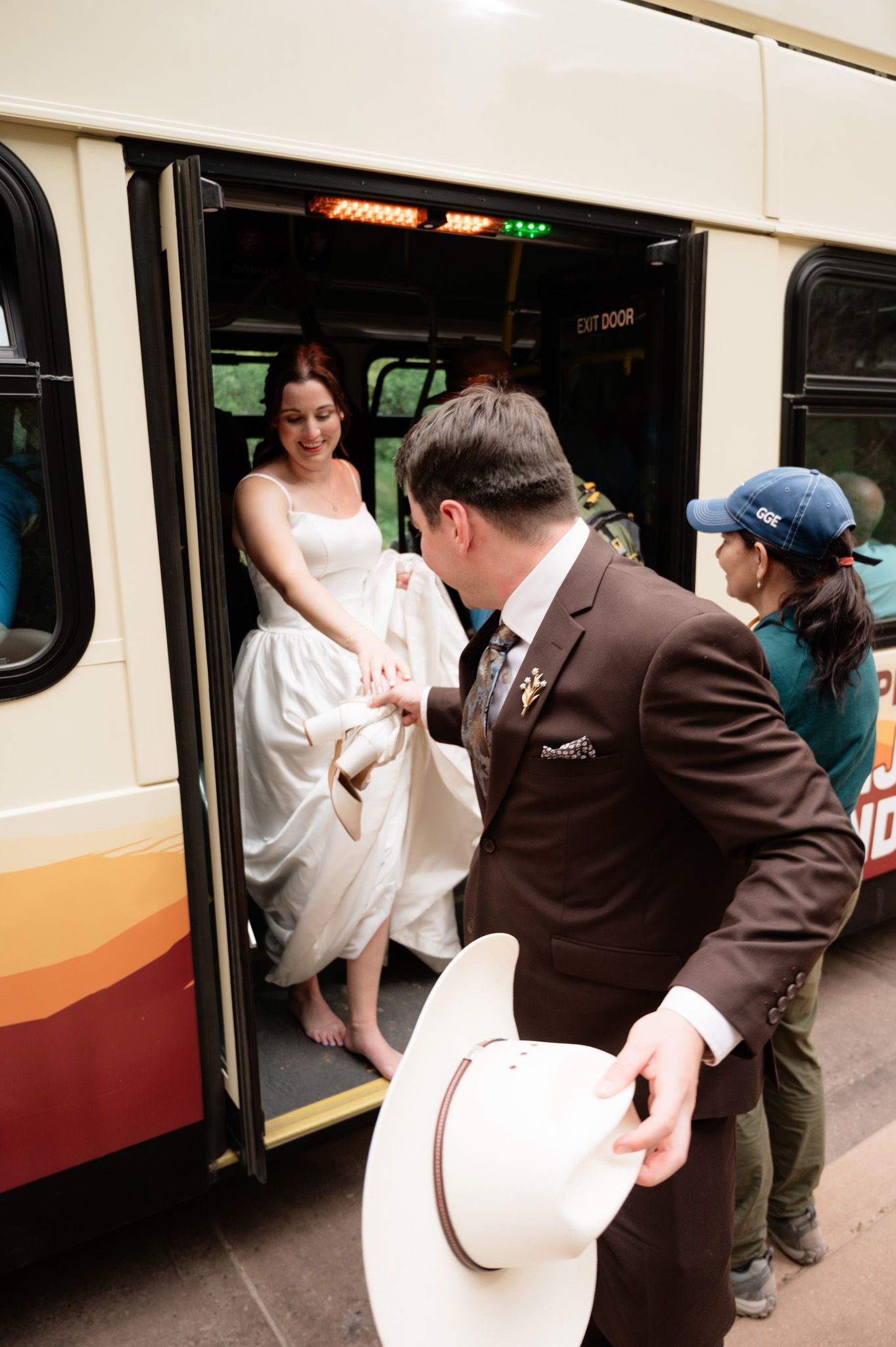 Groom helps bride in white wedding dress exit a Zion National Park shuttle bus while holding a cowboy hat.