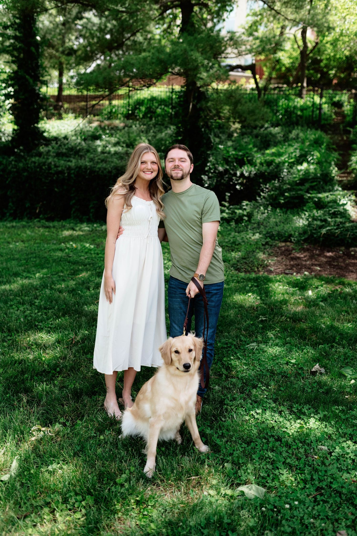A couple poses with their golden retriever in a lush green park setting.