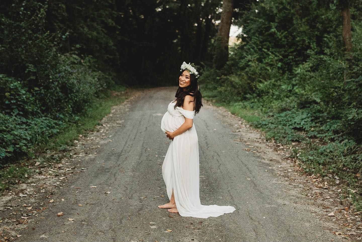 Artistic maternity portraits in flowing white dress on wooded path.