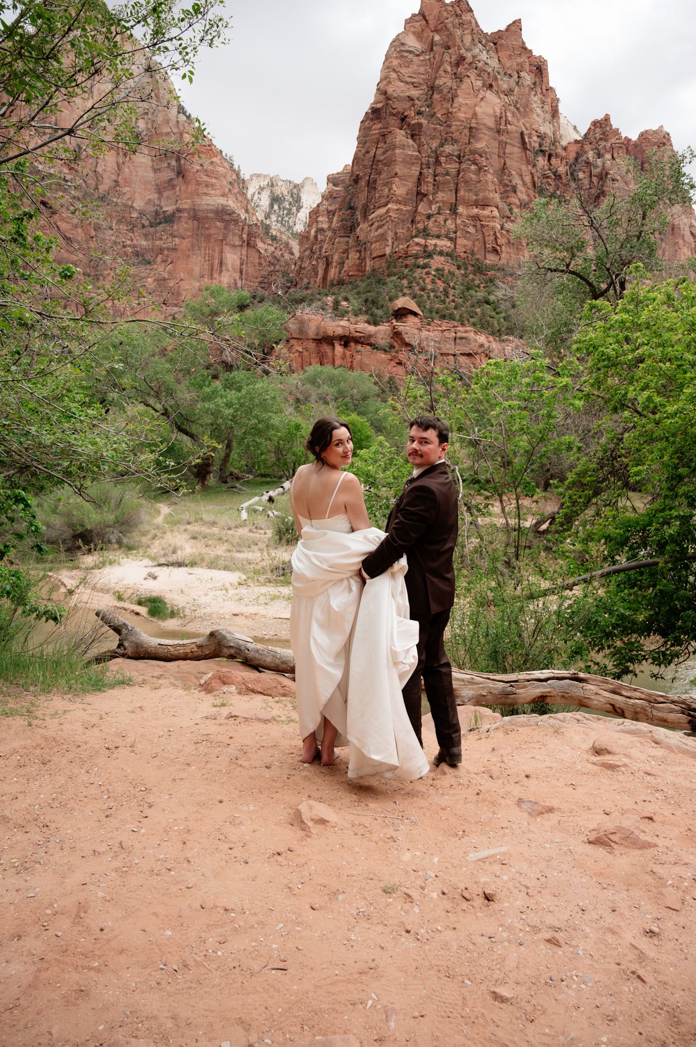 Bride and groom hold hands dancing at Zion National Park with red rock canyon formations and green trees behind them.