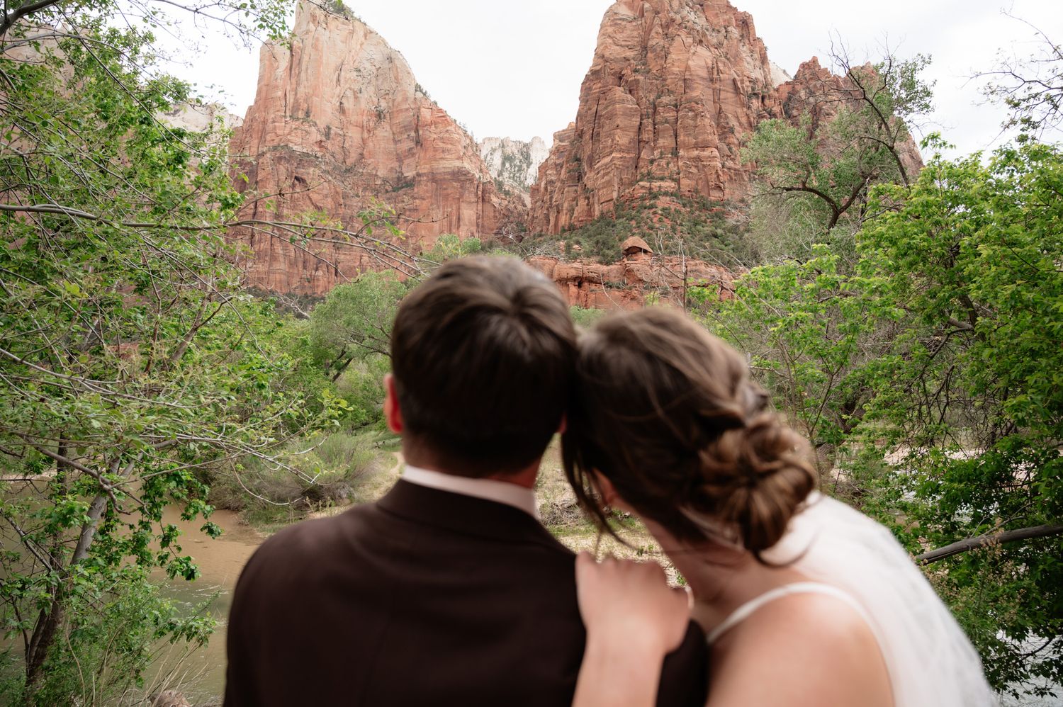 Bride and groom lean together gazing at red rock canyon cliffs surrounded by lush green trees at Zion National Park.