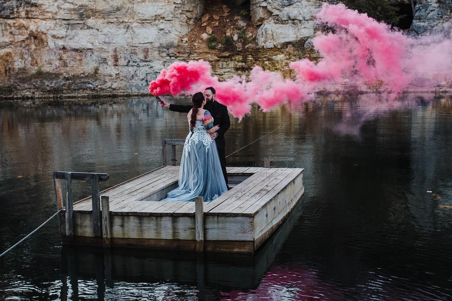 Wedding couple on dock with pink smoke bombs creating dreamy atmosphere over water.