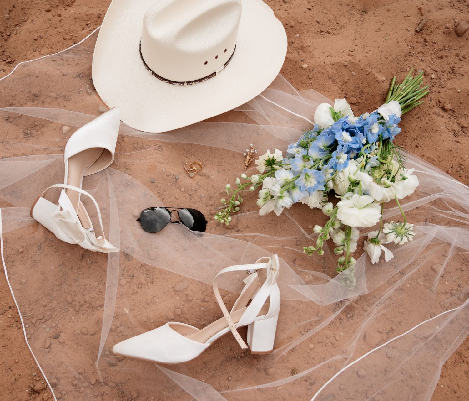 Bridal flat lay on sandy ground featuring white heels, cowboy hat, veil, sunglasses, and blue and white flower bouquet.