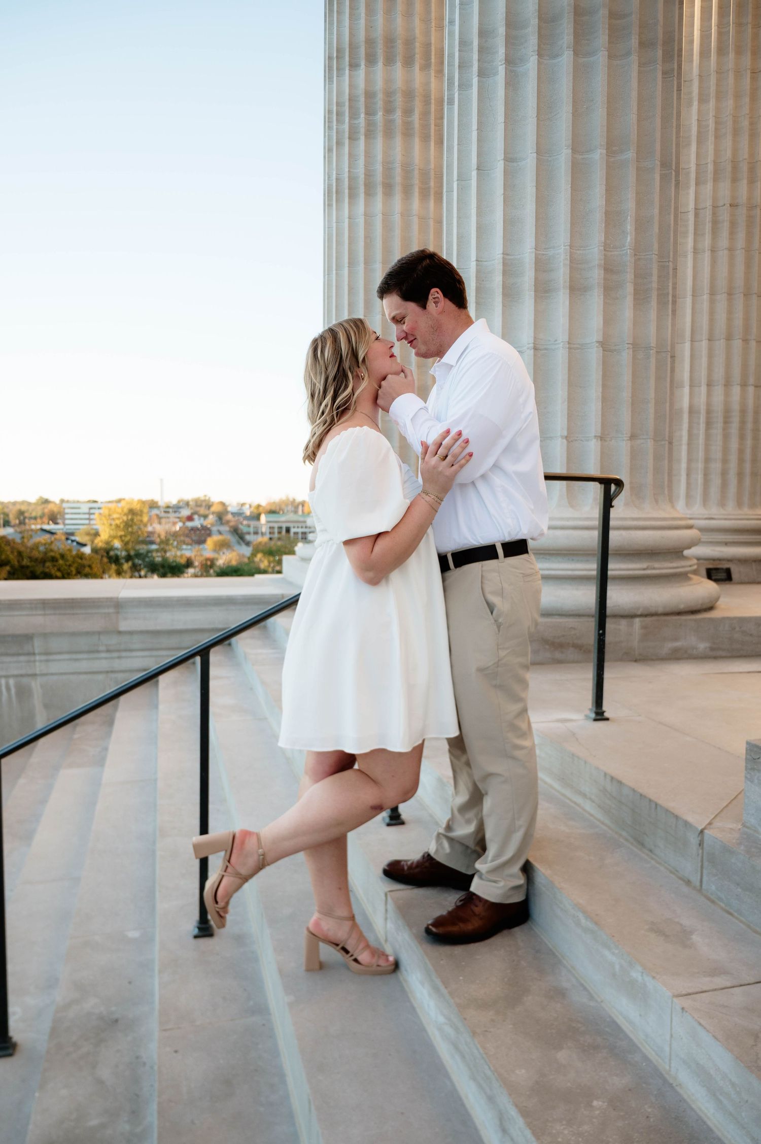 Couple sharing a romantic moment on stone steps near large columns, woman in white dress, man in white shirt and khakis.