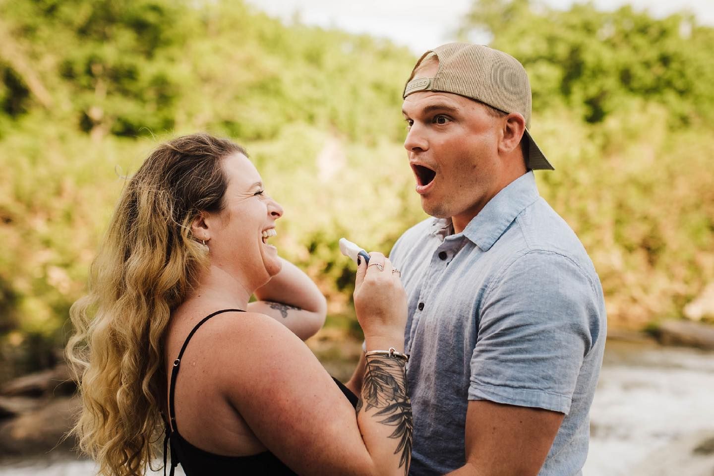 Playful couple laughing and embracing by a river during a summer photoshoot.
