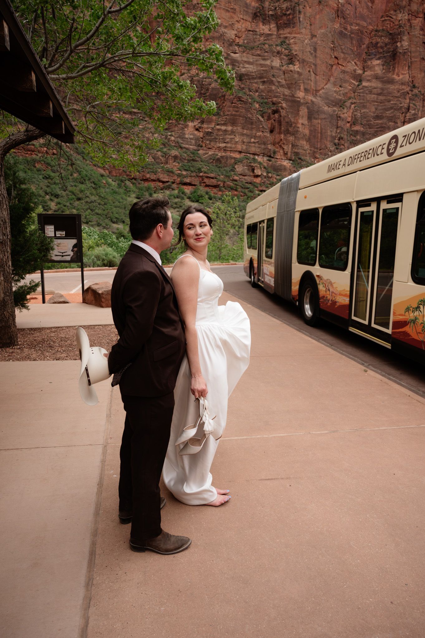 Bride in white gown and groom in dark suit stand together near a Zion National Park shuttle bus.