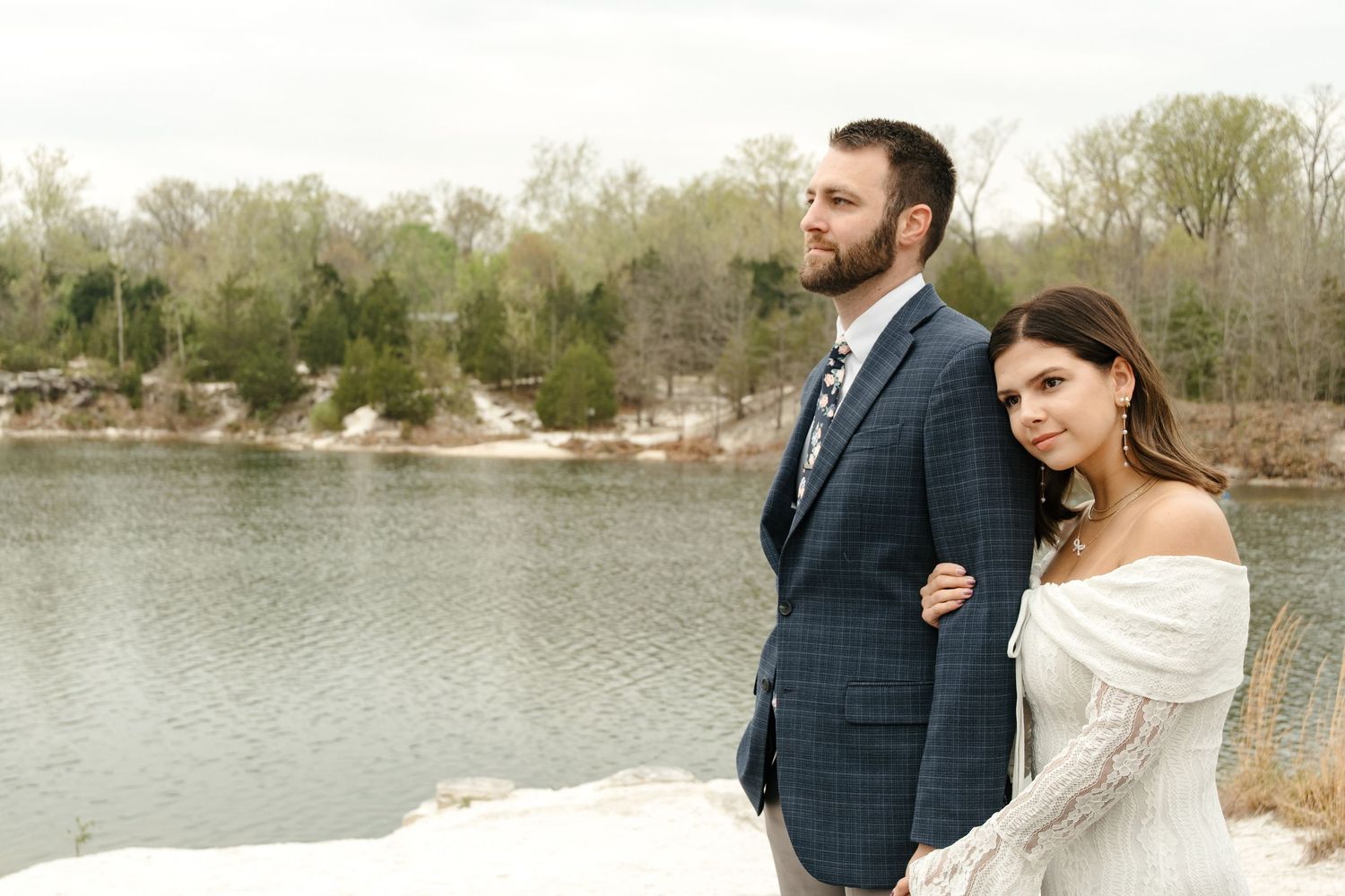 Bride and groom pose by a snowy riverbank with bare trees, woman in off-shoulder gown, man in blue plaid suit.