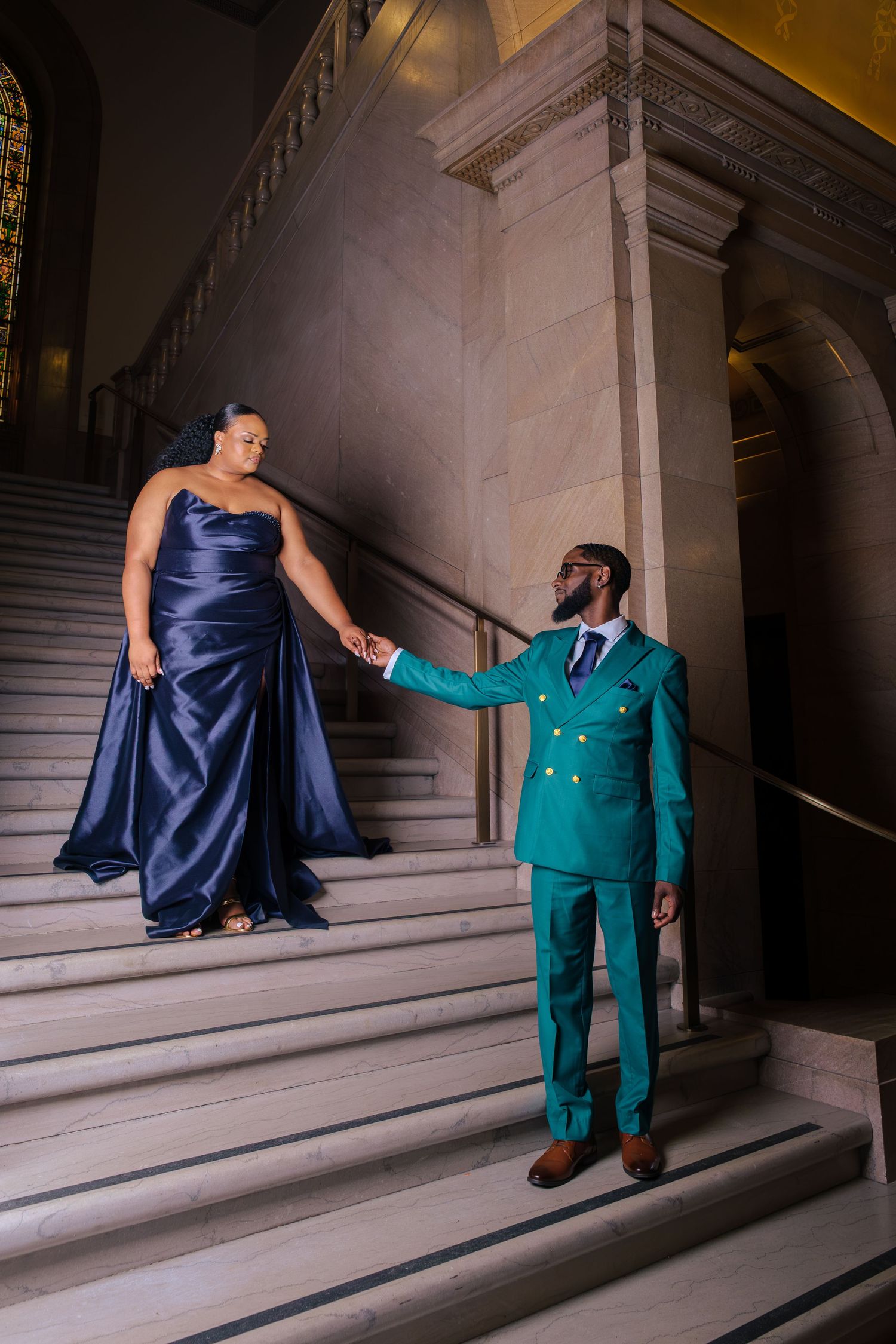 Couple in elegant formal wear on grand marble staircase, woman in navy gown, man in teal suit holding hands.