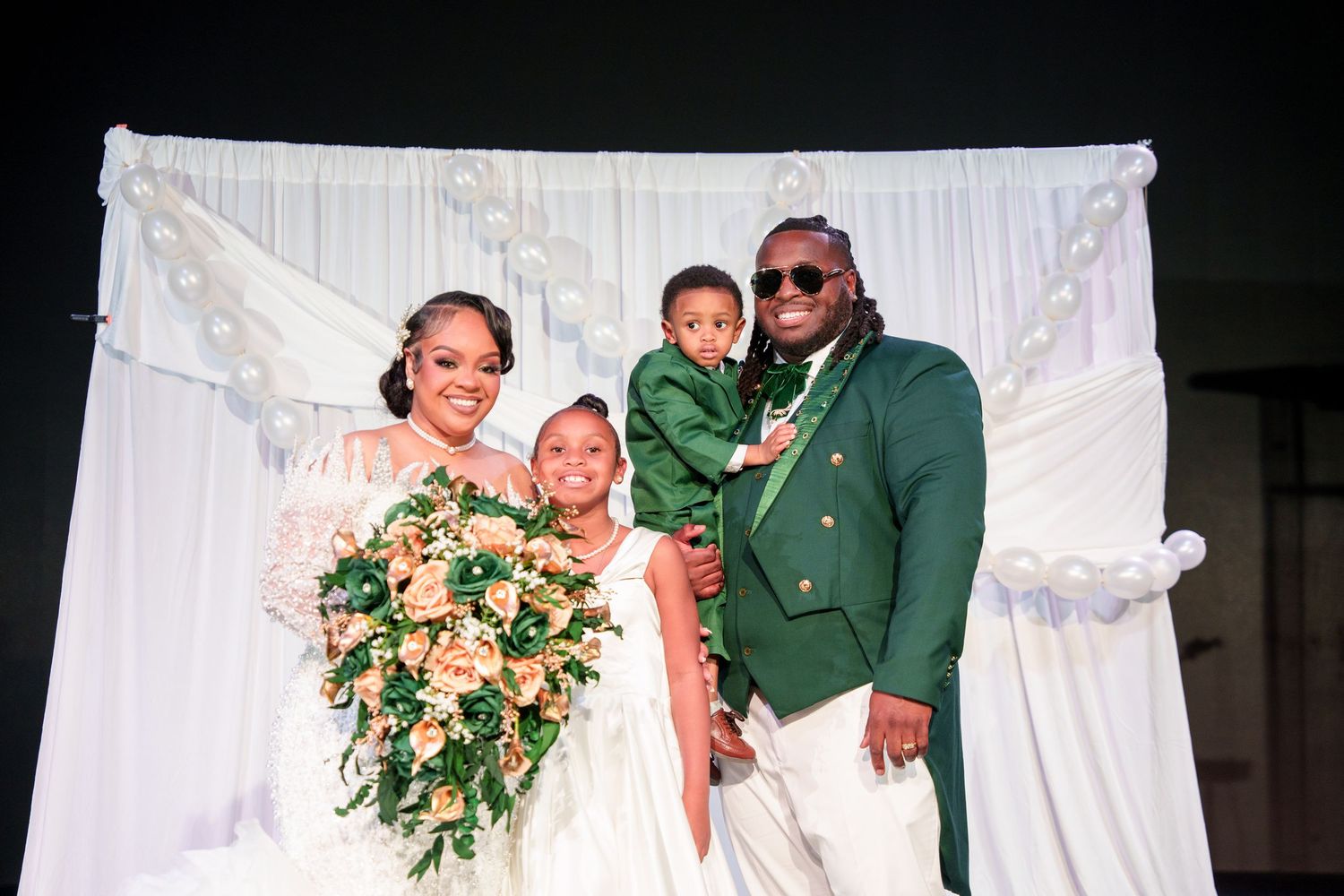 Wedding party poses against white backdrop with floral bouquets and green attire.