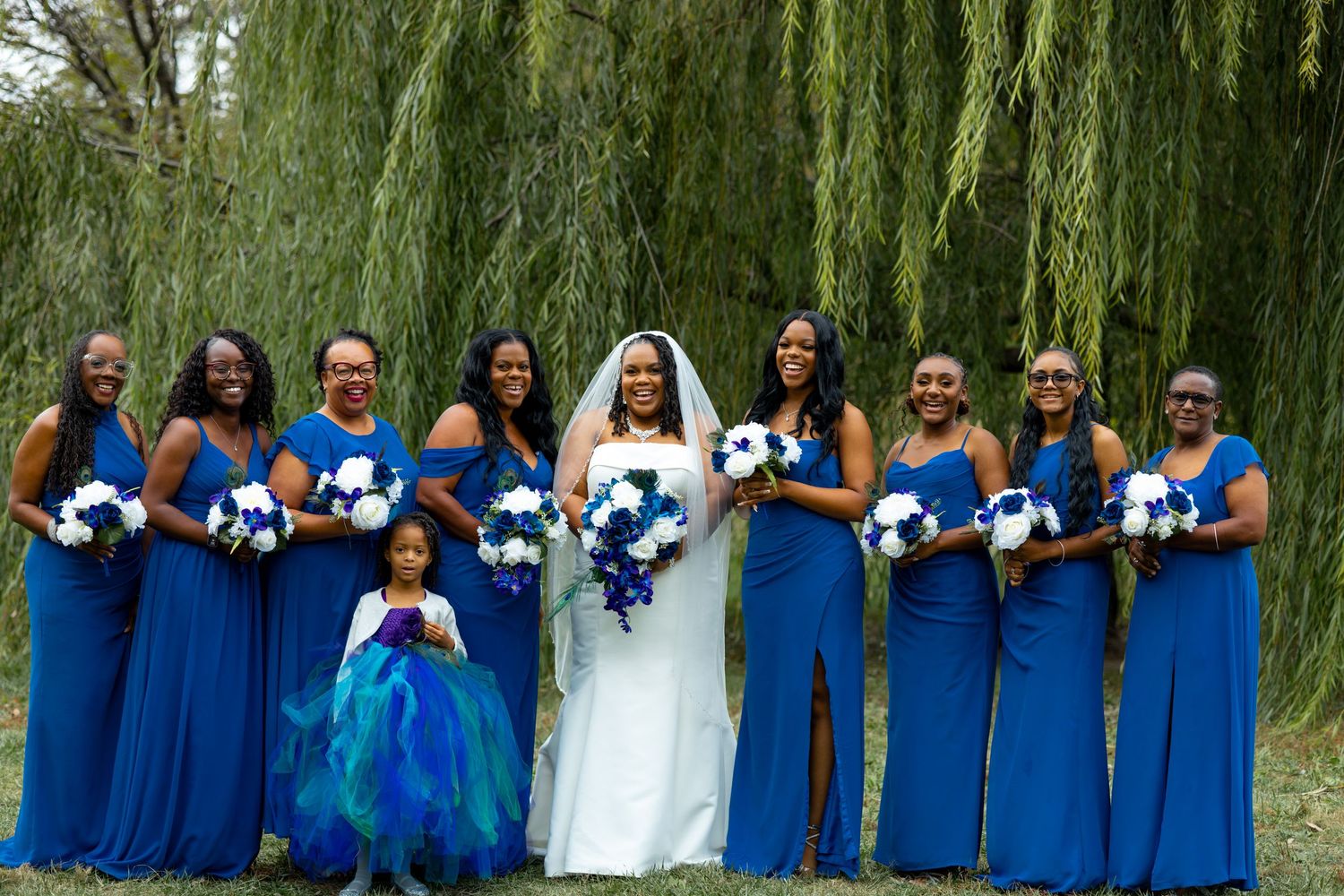 Wedding party in royal blue dresses poses under a weeping willow tree holding blue and white bouquets.