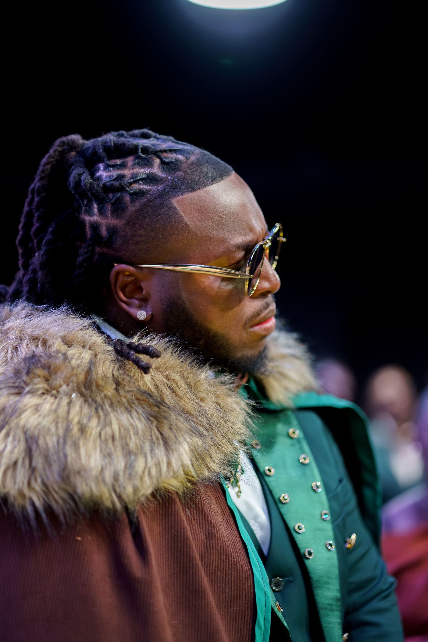 A person wearing sunglasses and a green jacket with fur trim collar is shown in profile against a dark background.