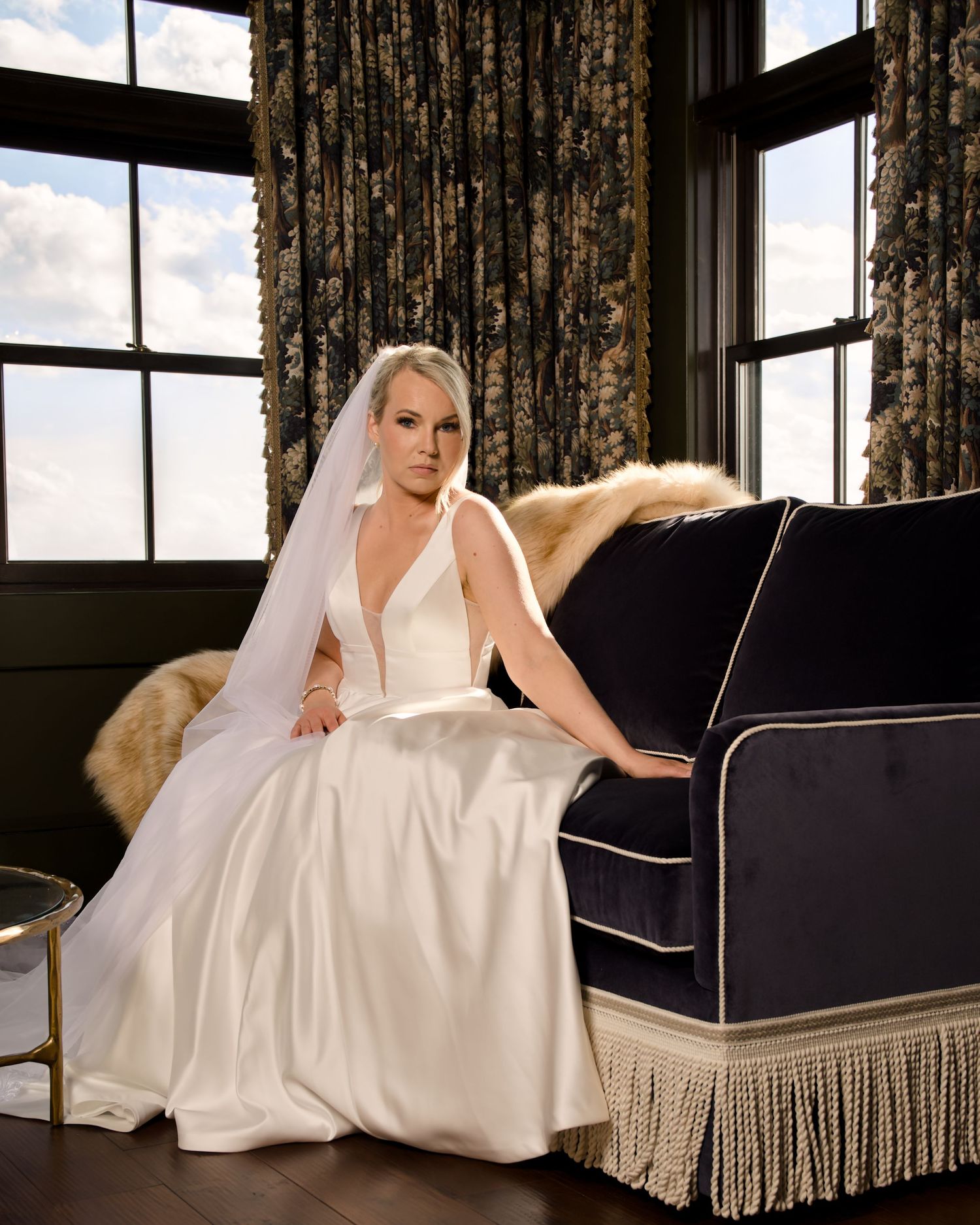 Bride in elegant white satin gown and veil seated on navy blue sofa in luxurious dark interior setting.
