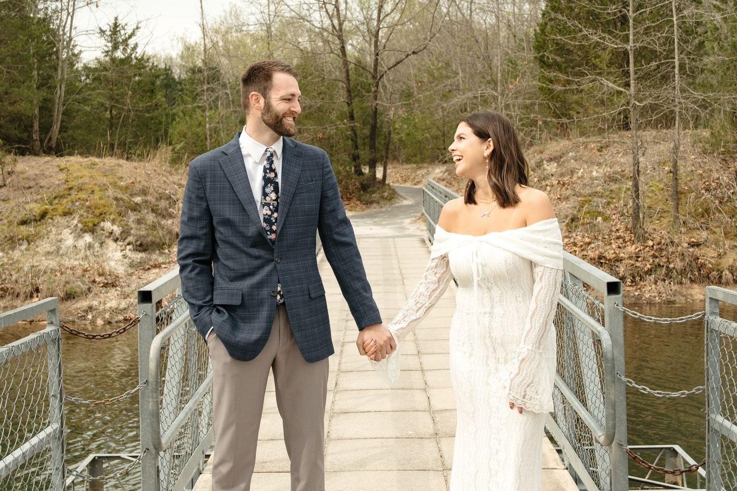 Bride and groom holding hands on a wooden bridge surrounded by trees, smiling at each other on their wedding day.