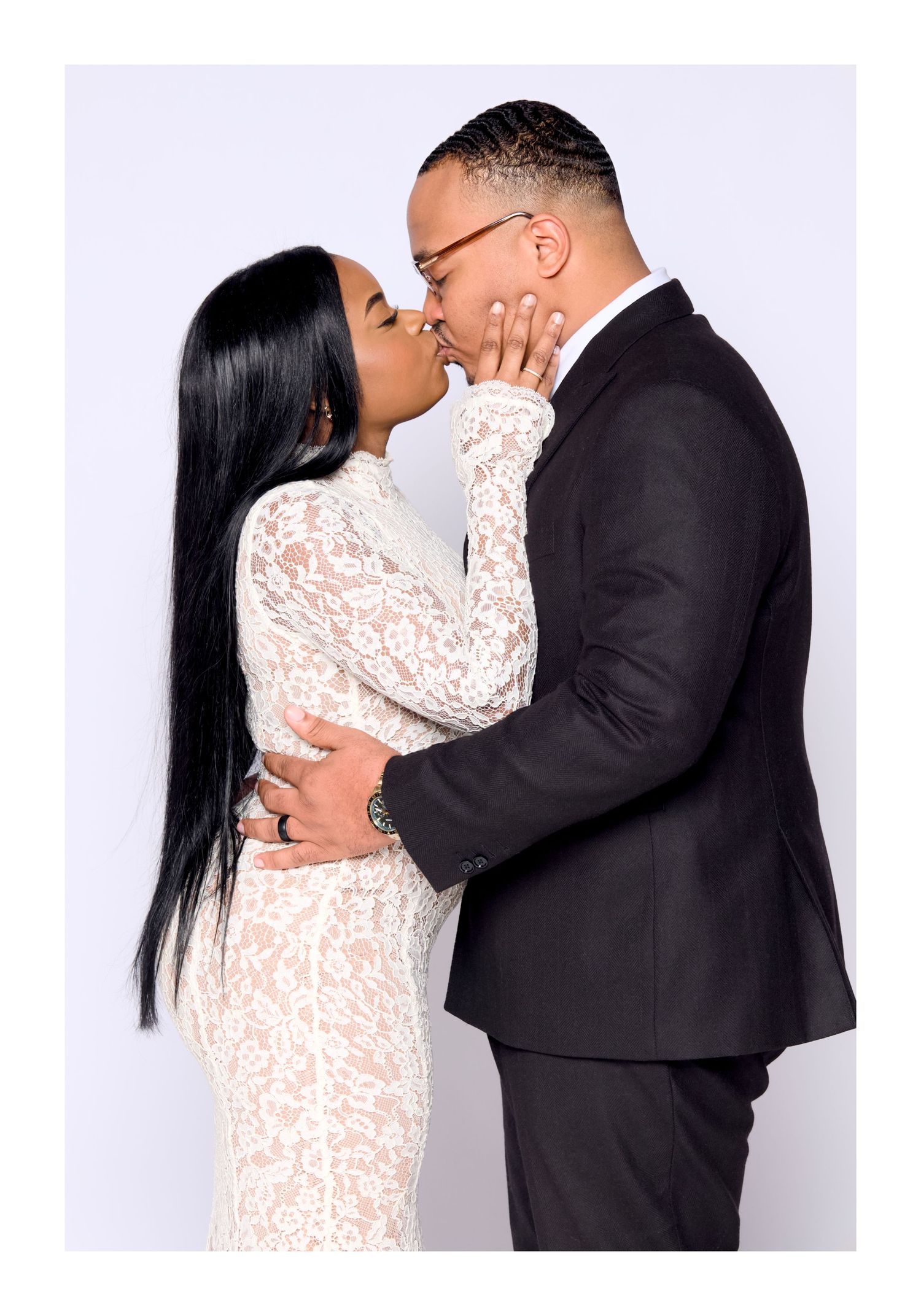 A couple shares a romantic kiss, woman in white lace dress and man in black suit, against a white background.