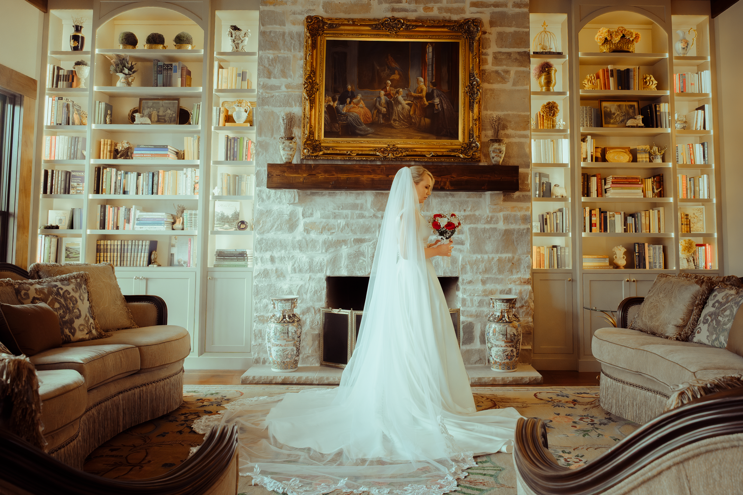 Bride in white gown and veil stands in elegant library with stone fireplace, bookshelves, and classic painting.