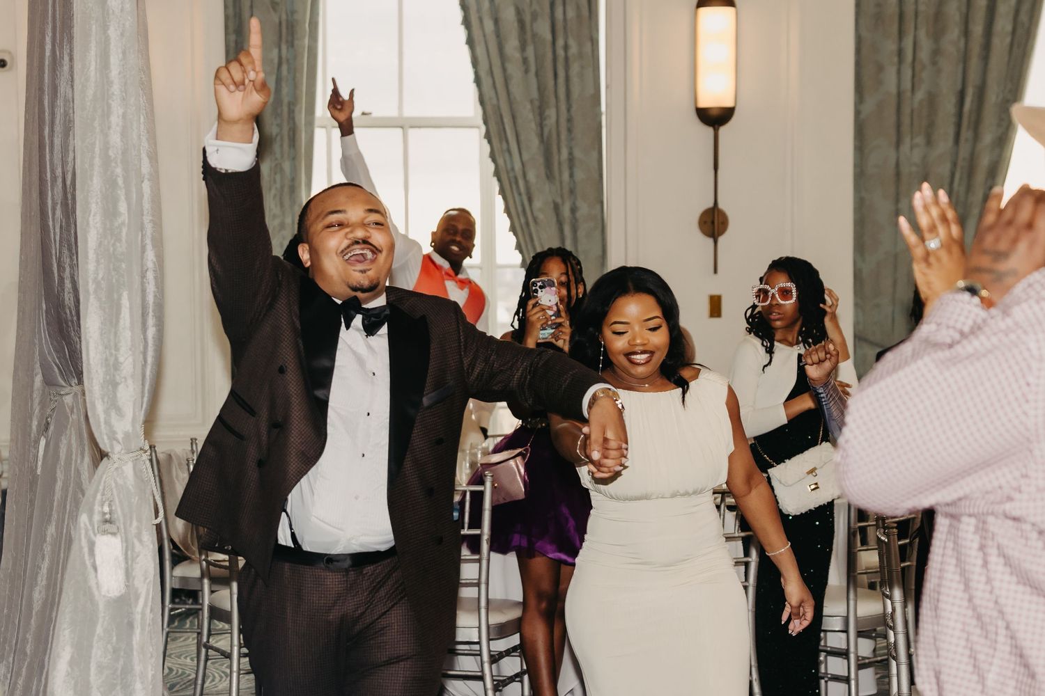 Wedding party celebrates with joyful dancing and raised hands in an elegant ballroom with white drapes.