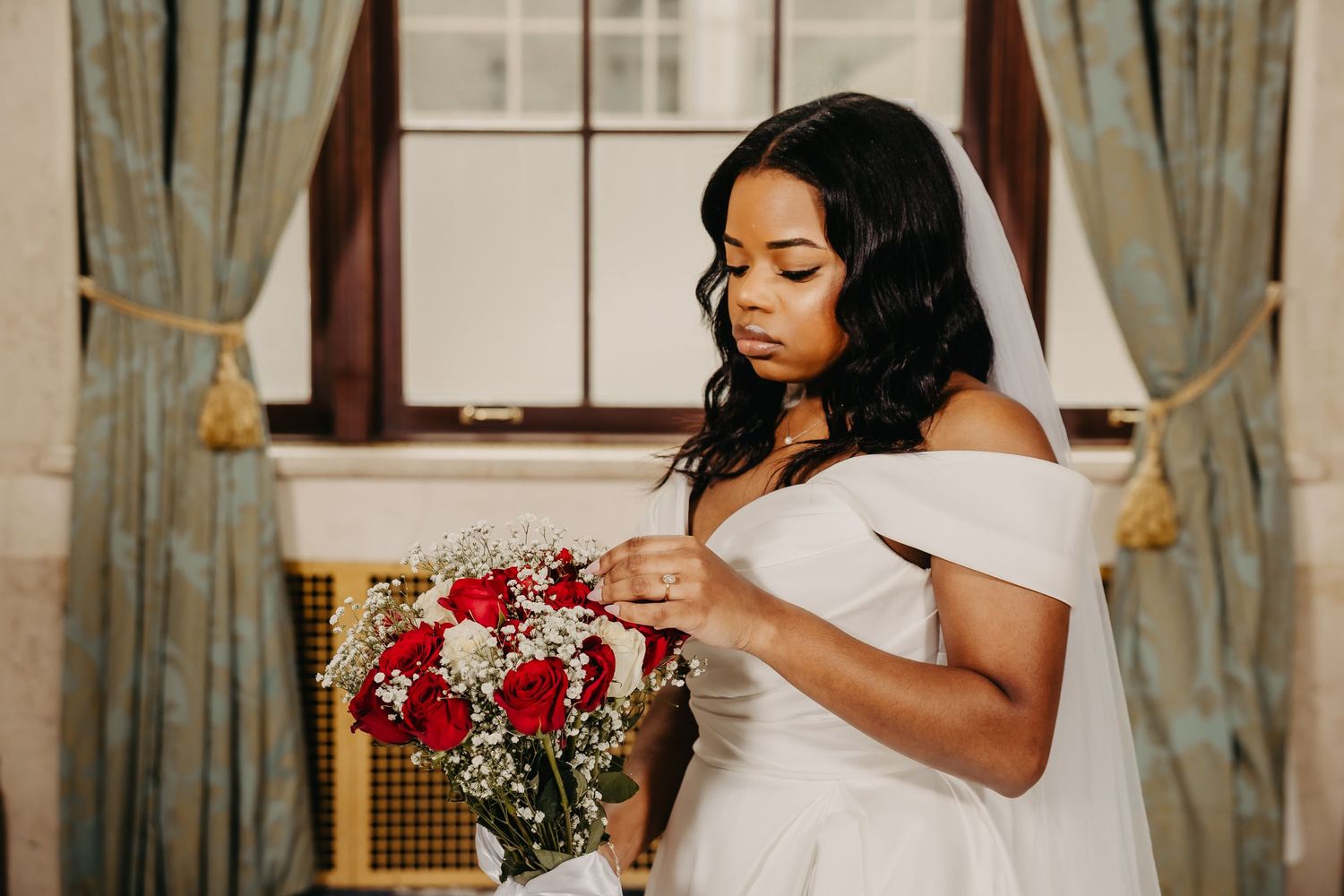 Bride in white gown holds red bouquet during elegant indoor wedding photoshoot.