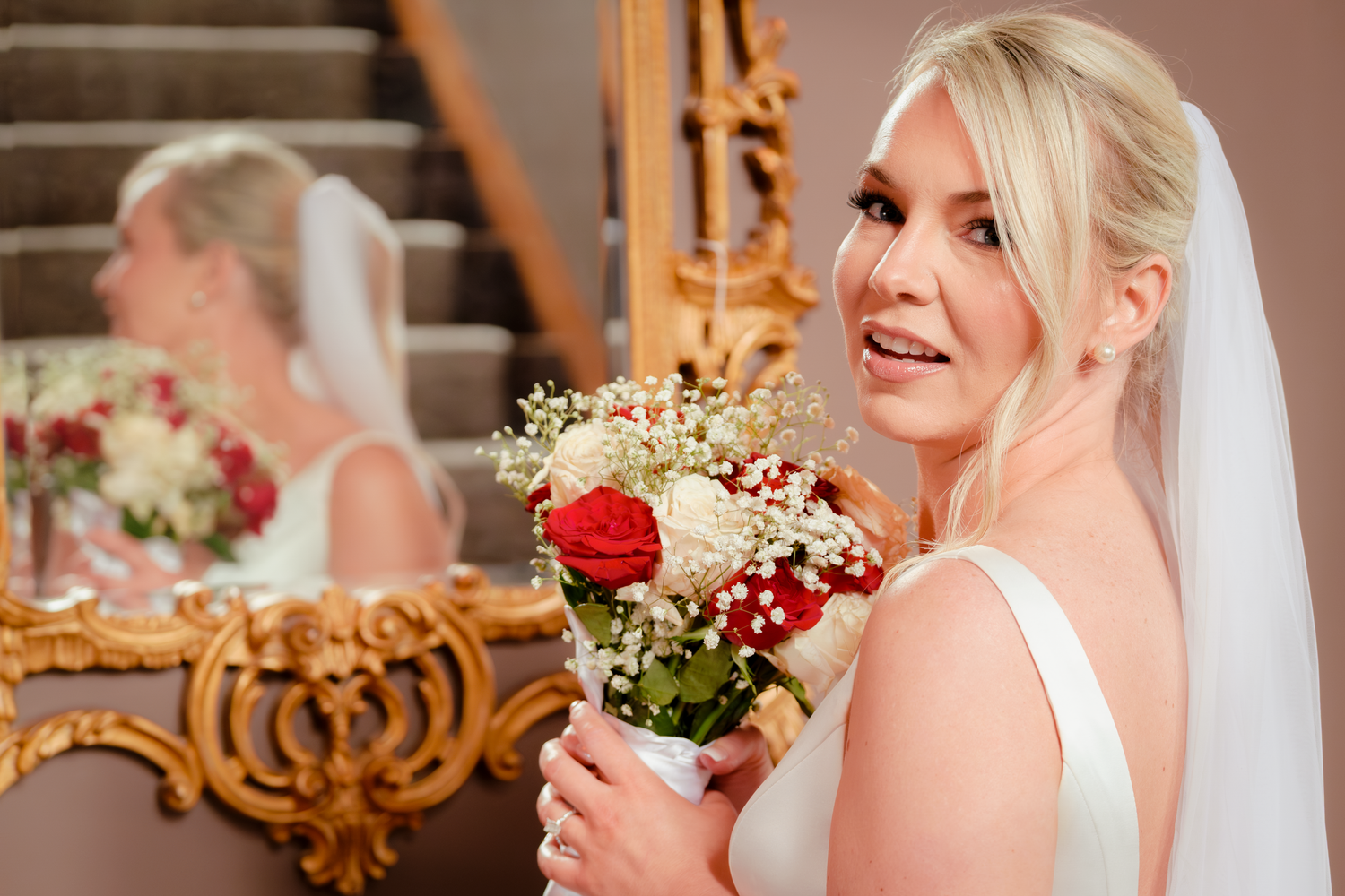Blonde bride in white gown holds red and white bouquet, smiling near ornate gold mirror.
