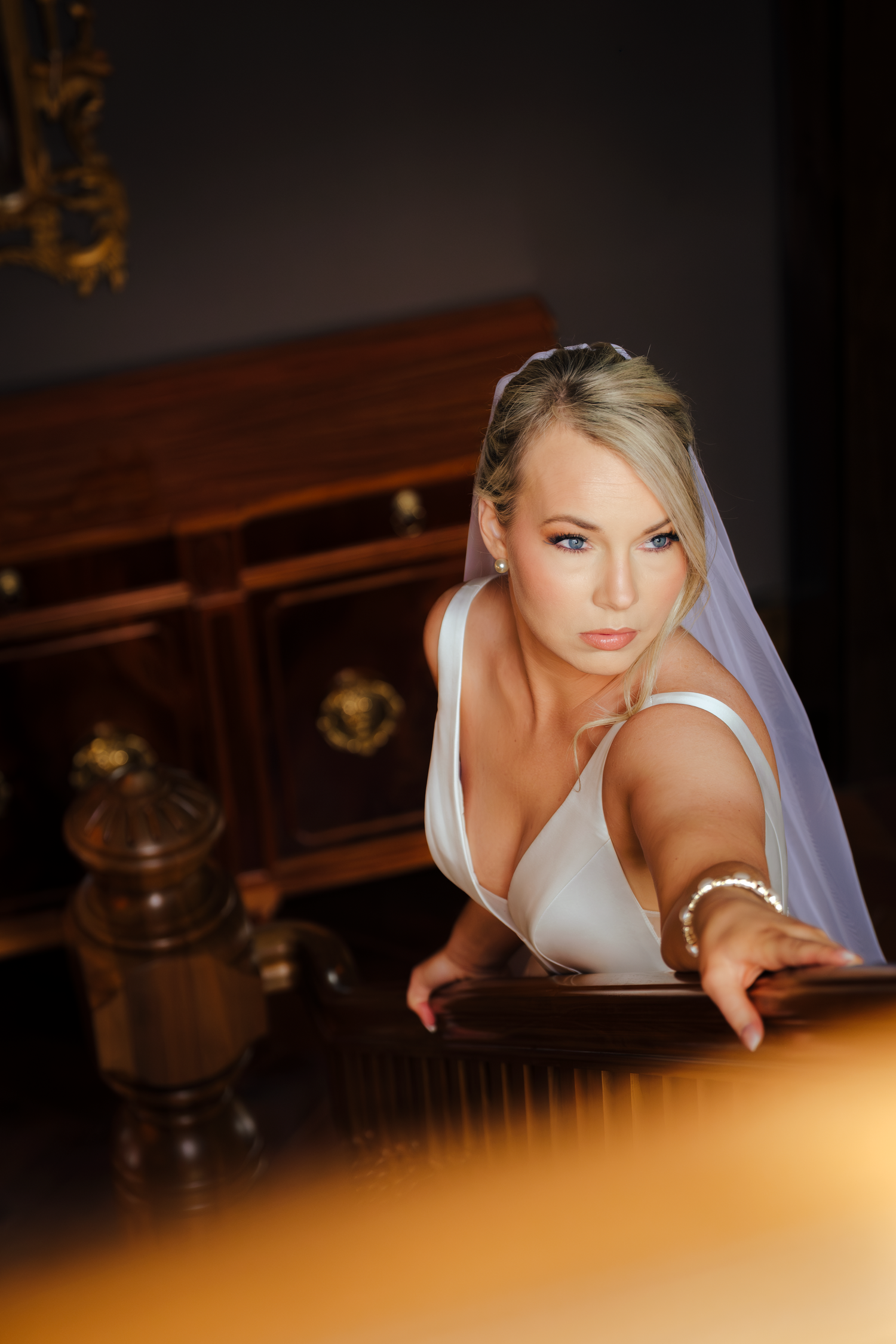 Blonde bride in white gown and veil leaning over a dark wooden staircase railing in an elegant interior.
