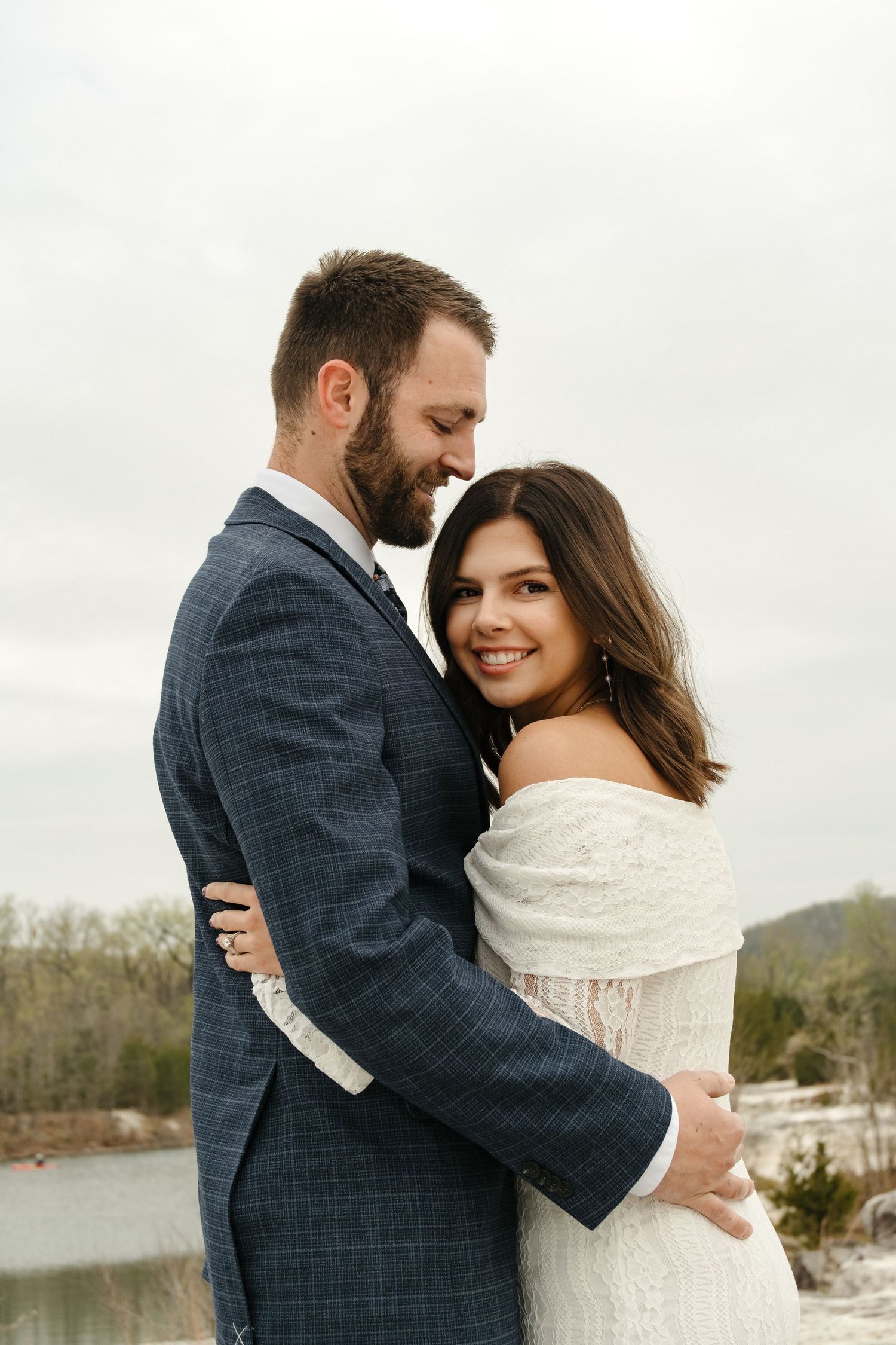A couple in wedding attire embraces outdoors, the bride in white and groom in a blue plaid suit, smiling near a winter lake.