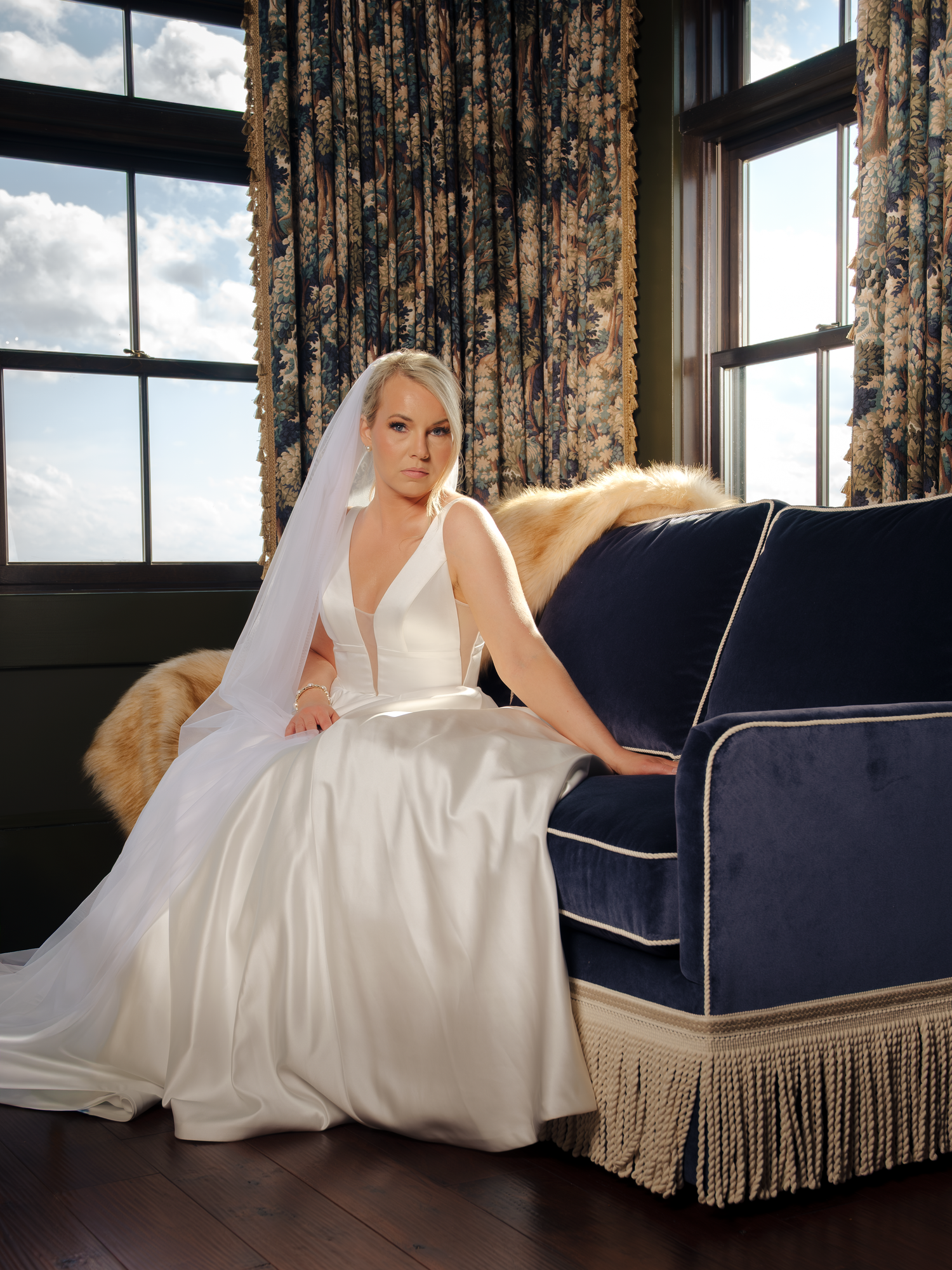Bride in white gown and veil seated beside a navy blue sofa with gold trim, posed elegantly near floral curtains.