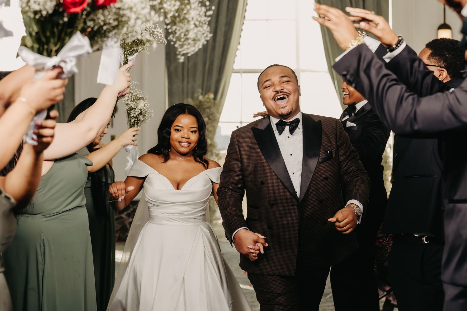 Wedding celebration with guests in formal attire against white drapes and floral decorations.