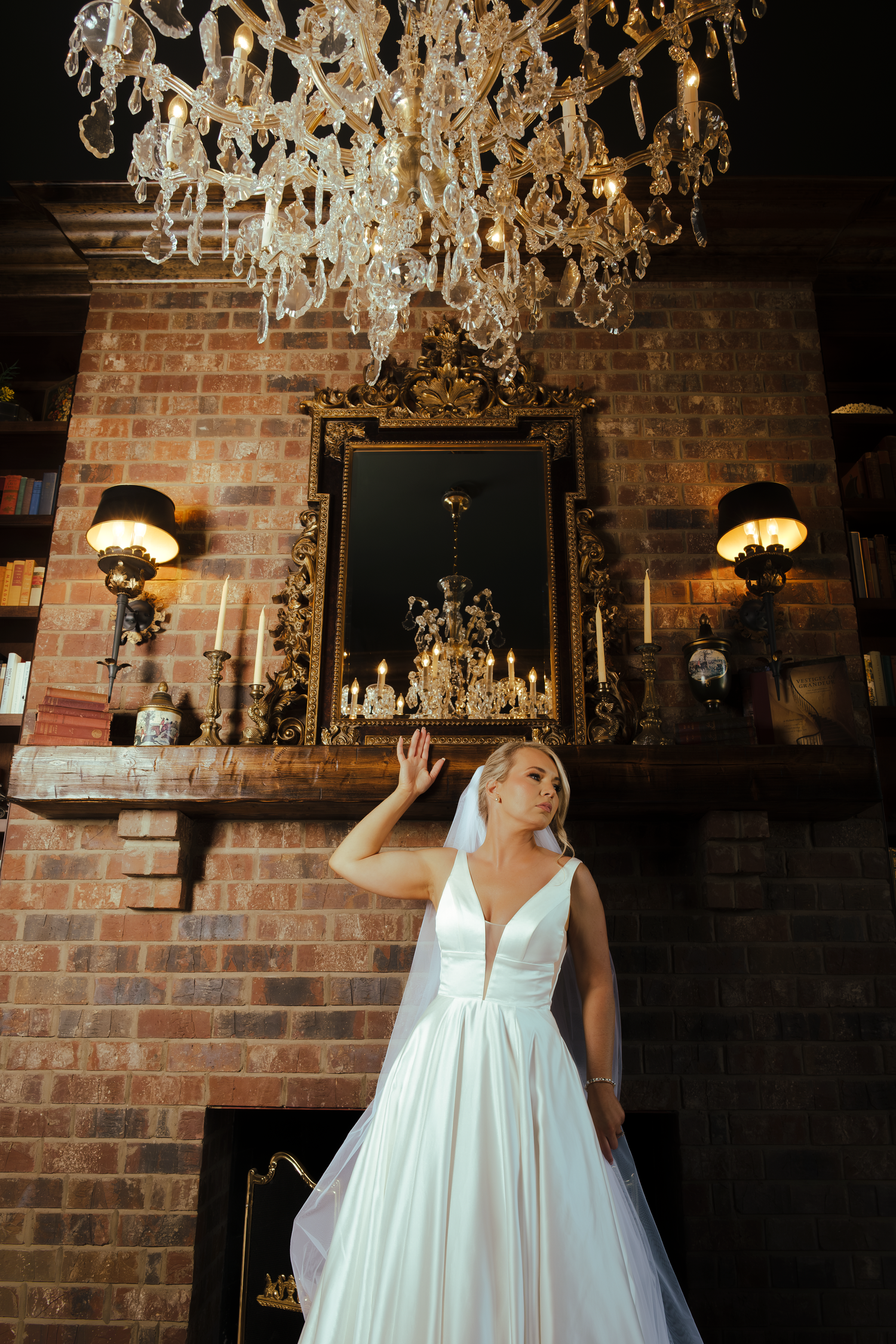 Bride in white ball gown poses under crystal chandelier near ornate fireplace in elegant brick-walled room.