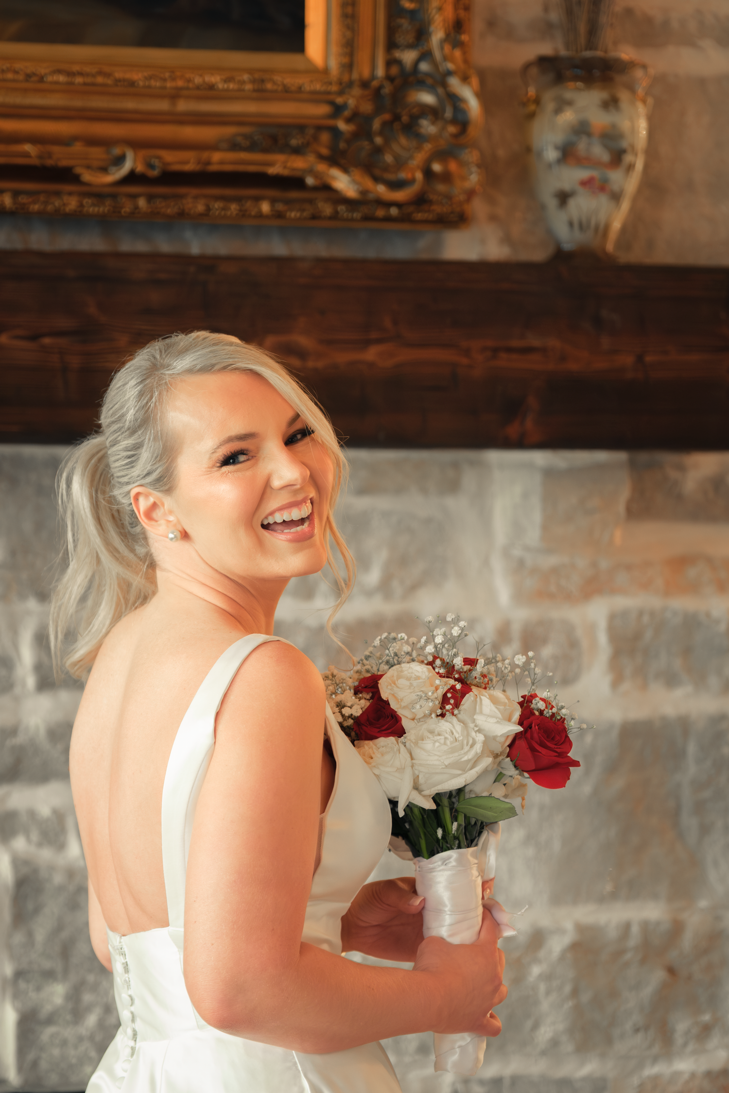 Smiling bride in white gown holding red and white bouquet standing in front of stone fireplace.
