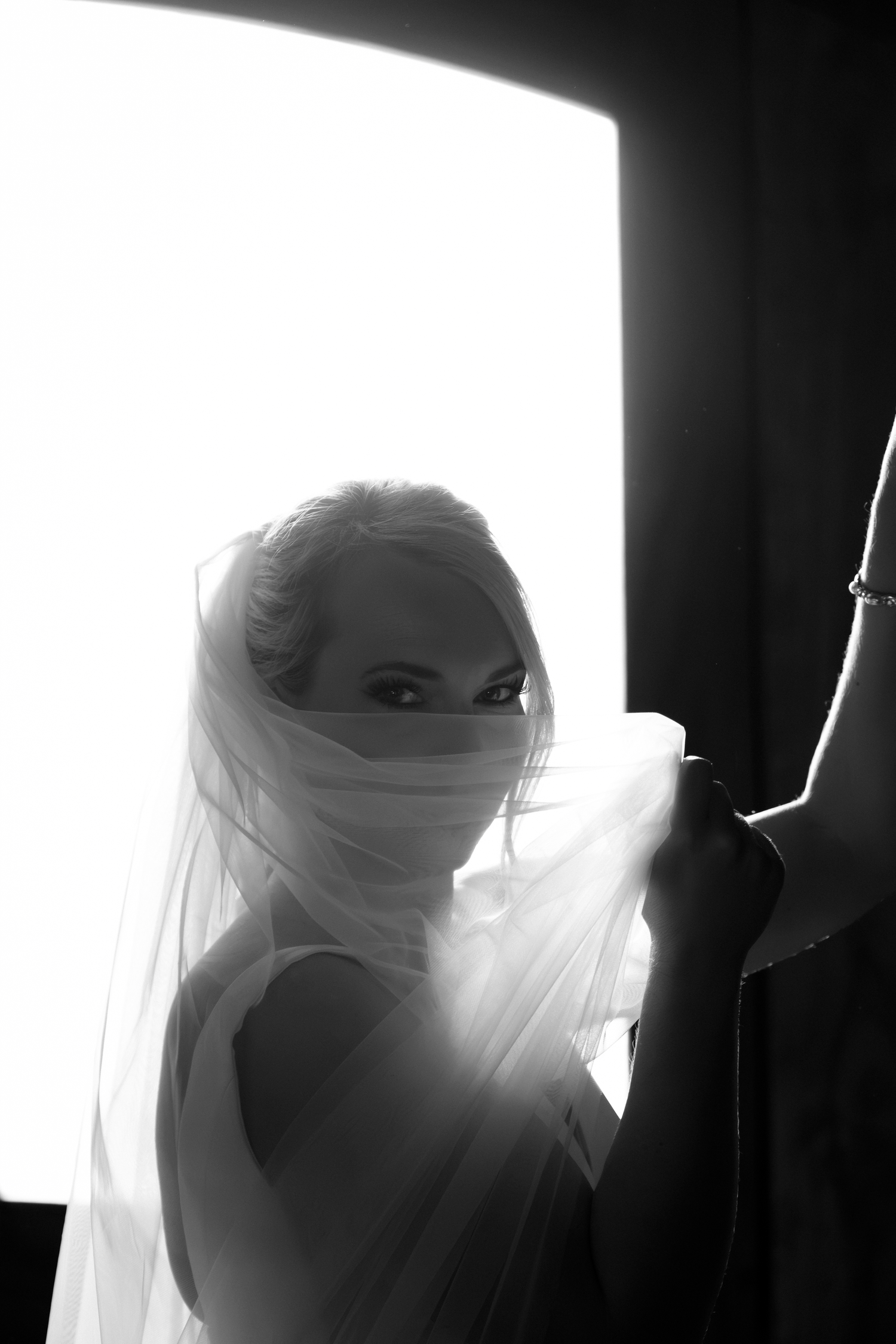 Black and white bridal portrait of a blonde bride with veil draped over her face, backlit by a bright window.