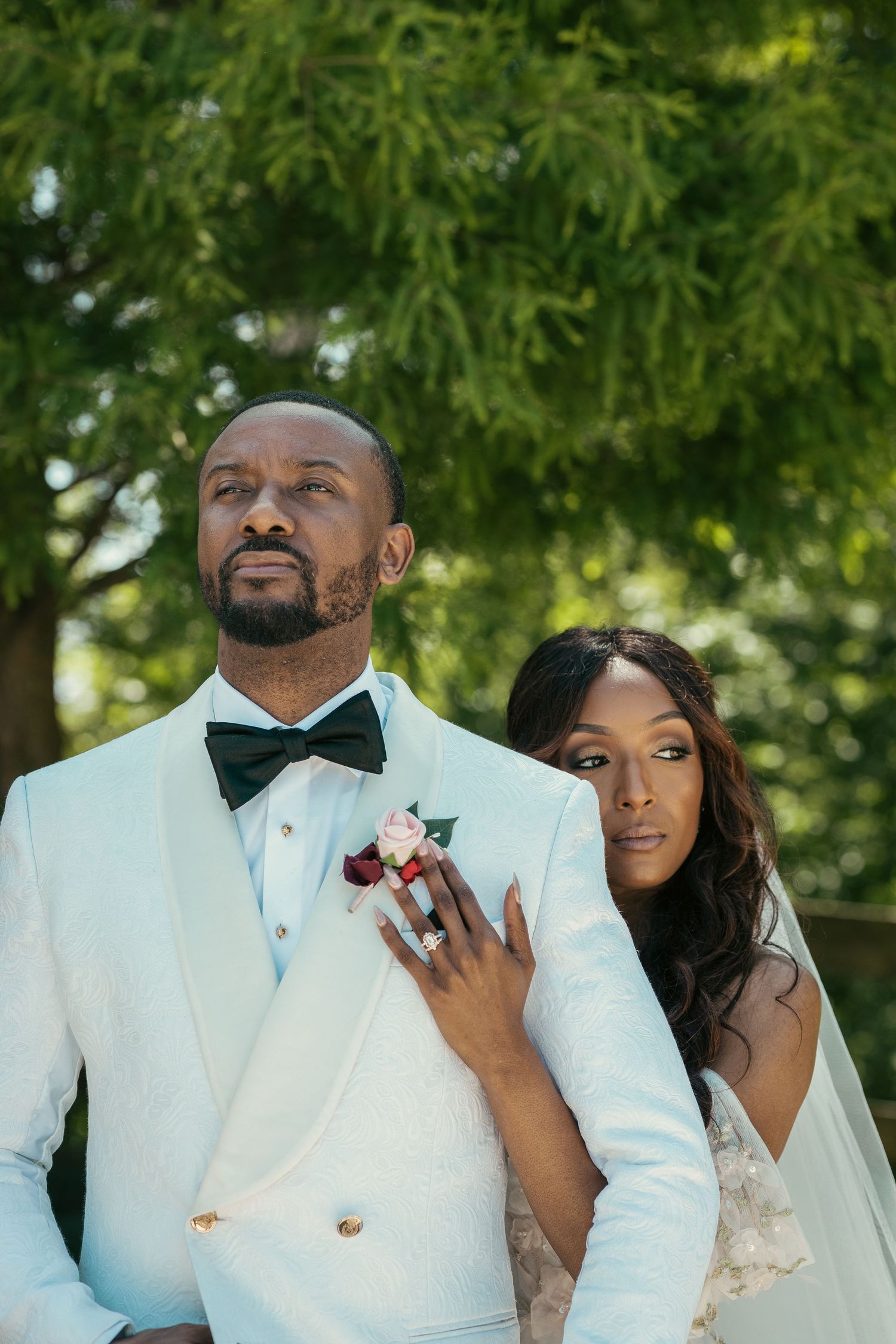 Wedding couple in white and cream formal attire posing together outdoors under green foliage.