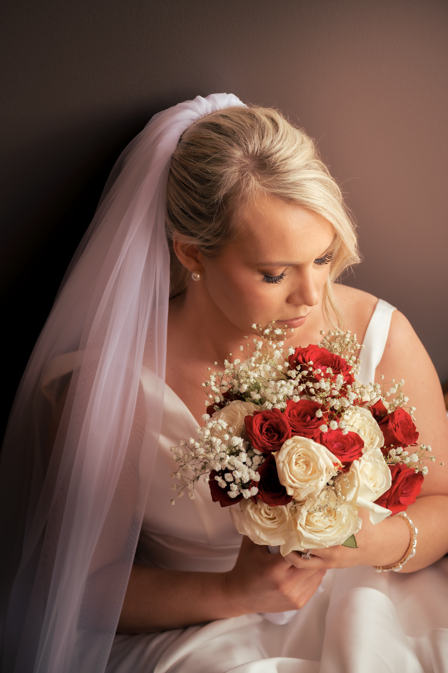 Blonde bride in white gown and veil gazes down at red and white rose bouquet with baby's breath.