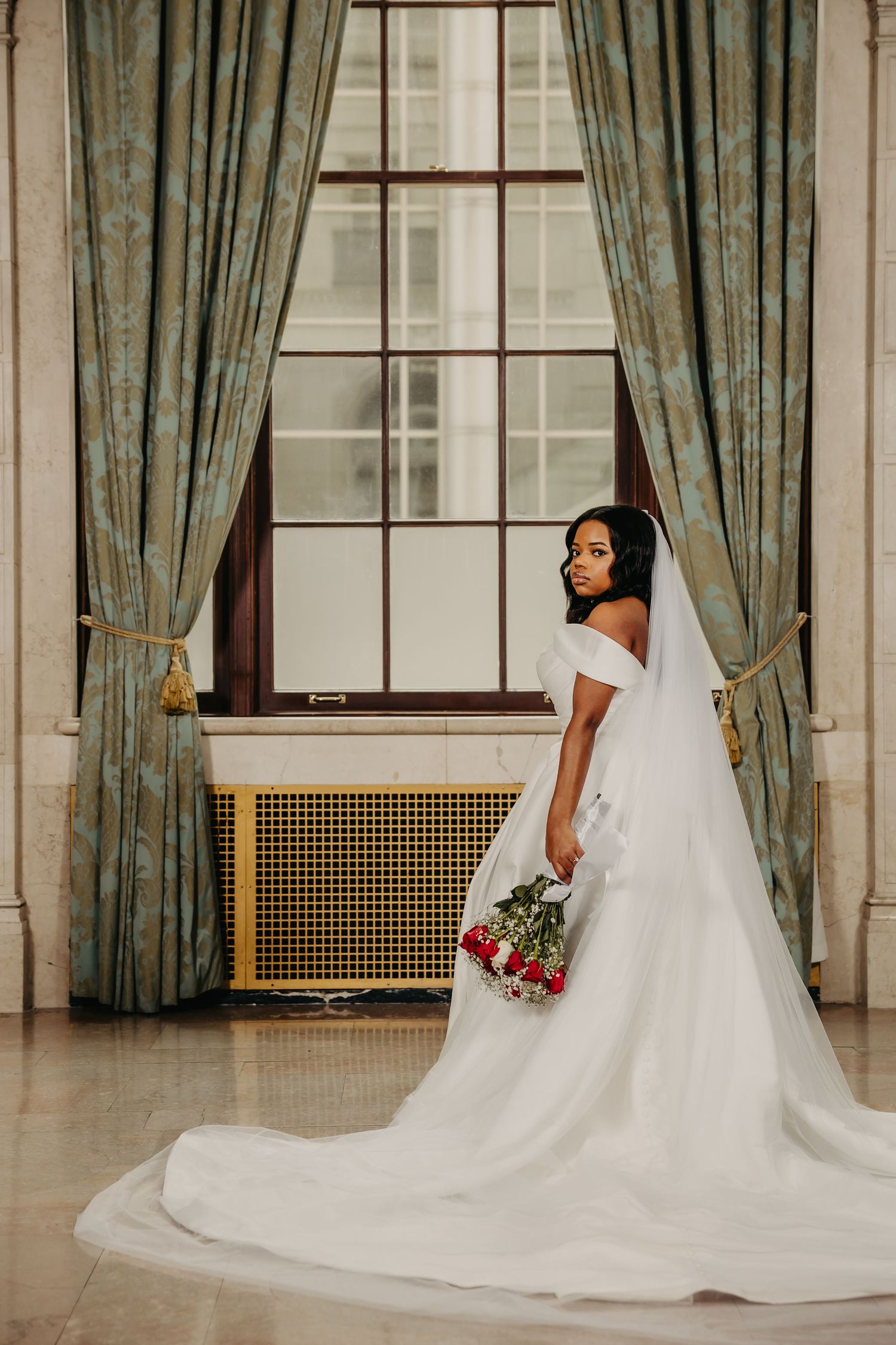 A bride in an elegant white wedding gown with long train poses by a window with sage green curtains holding a bouquet.